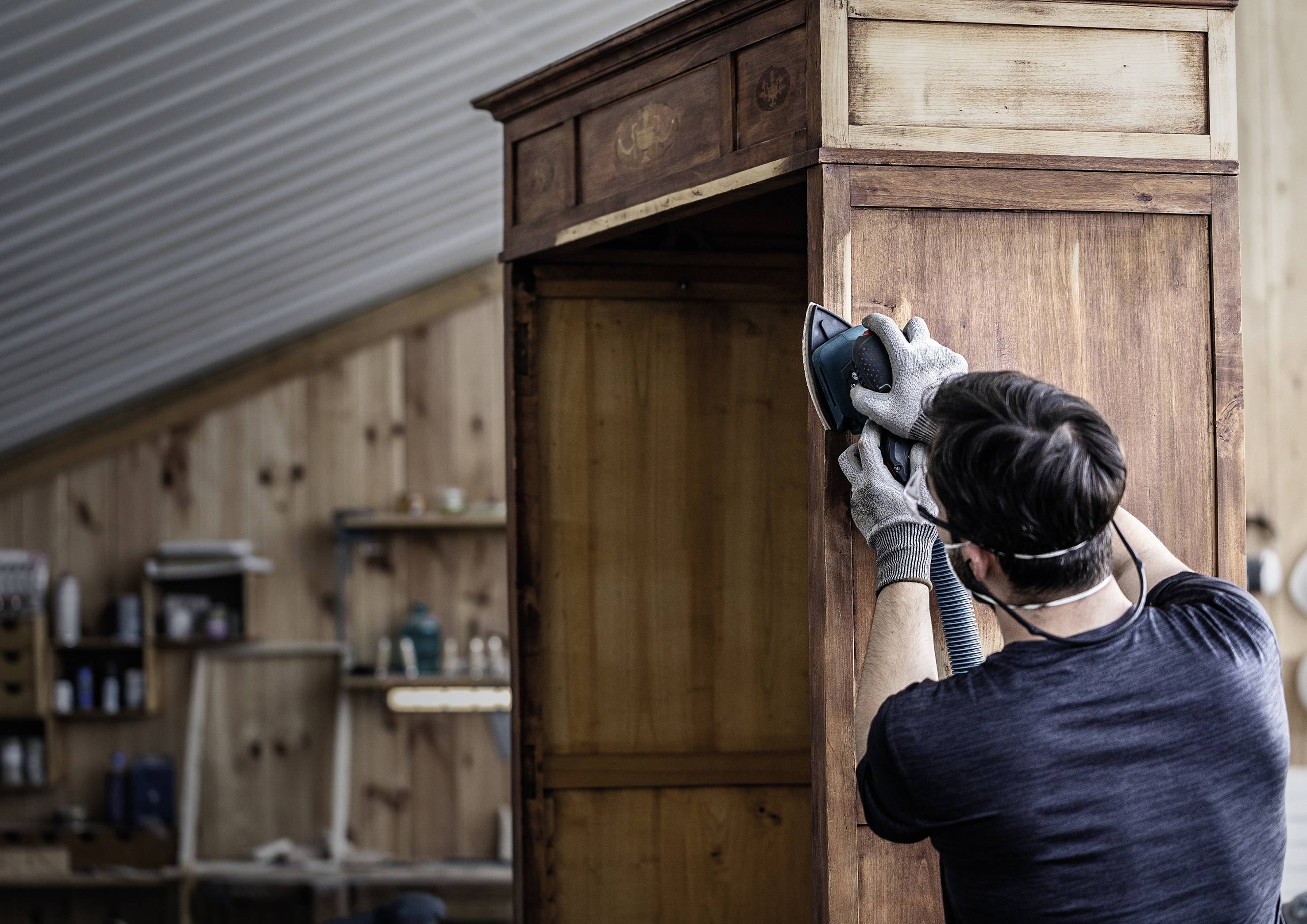 Eine Person bearbeitet ein Holzschrank mit einem Schleifgerät in einer Werkstatt. Der Raum hat holzverkleidete Wände und Regale im Hintergrund.