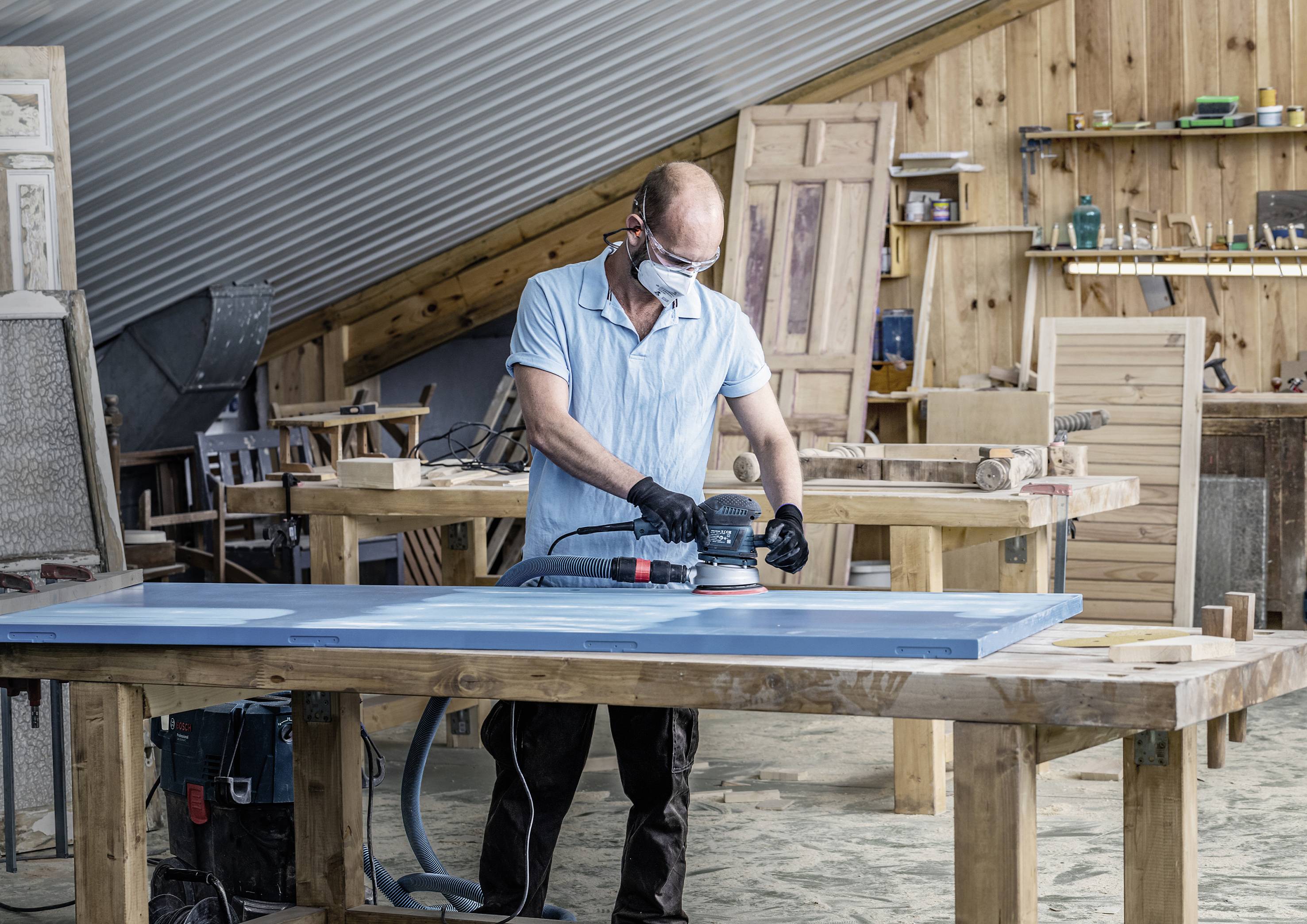 Ein Mann mit Schutzbrille und Maske schleift eine Holzplatte in einer Werkstatt mit Holzwänden und Handwerkswerkzeugen.