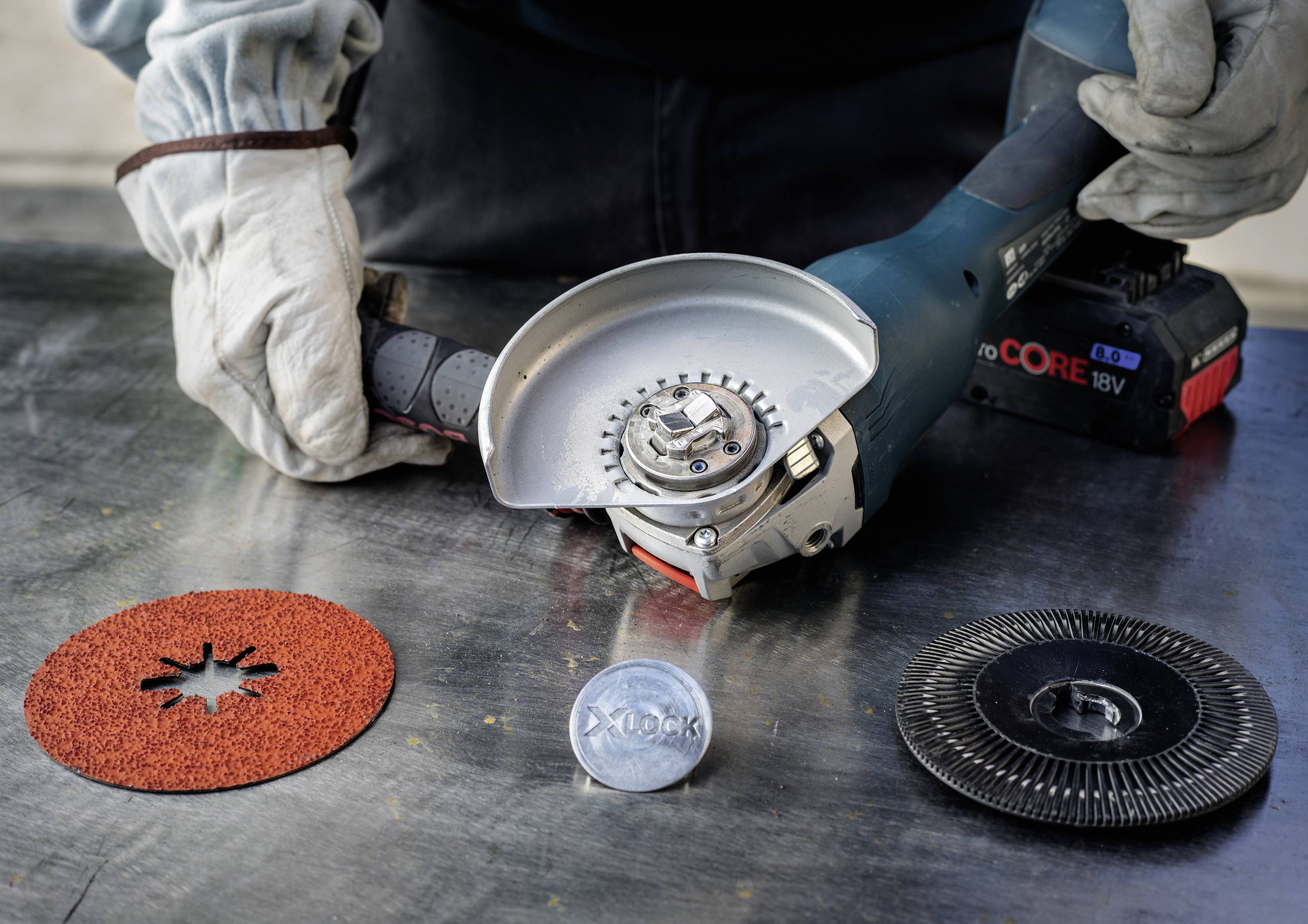 Eine Person trägt Schutzhandschuhe und hält einen Winkelschleifer über einem Metalltisch. Daneben liegen Schleifscheiben.