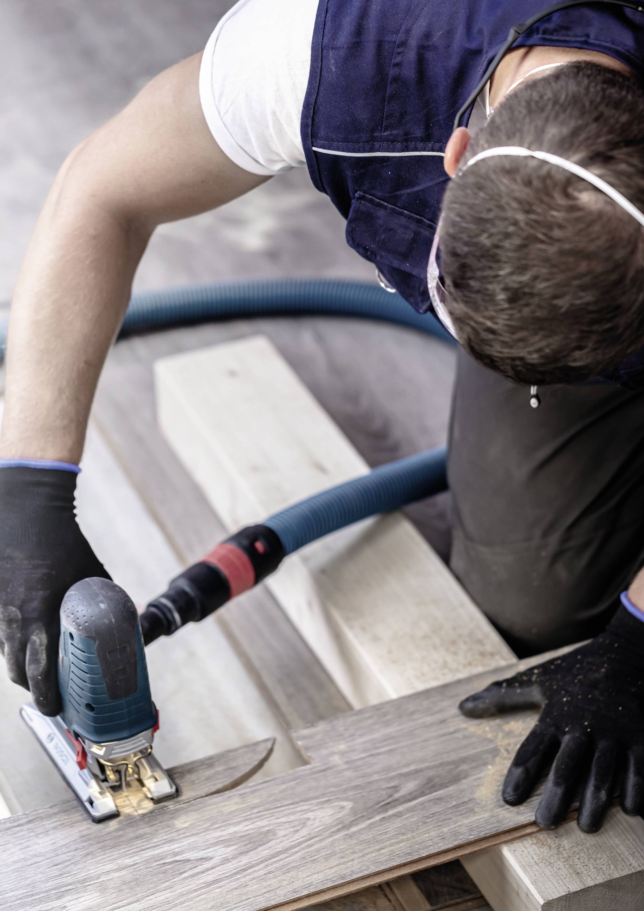 Ein Mann mit Schutzmaske benutzt eine Handkreissäge, um ein Holzbrett zu schneiden. Holzspäne liegen auf dem Boden.