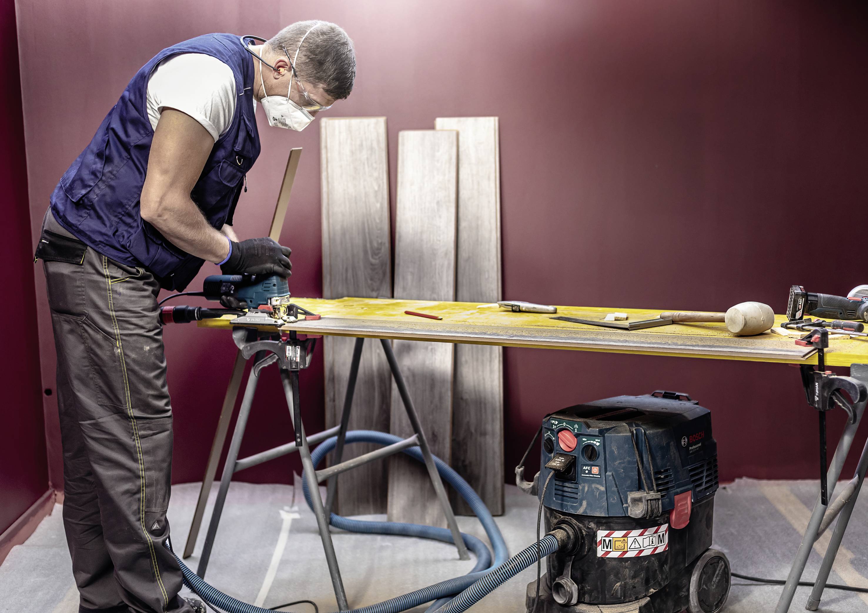 Ein Mann mit Schutzmaske arbeitet in einer Holzwerkstatt. Auf dem Tisch liegen Werkzeuge und Holzstücke. Ein Staubsauger ist neben ihm.