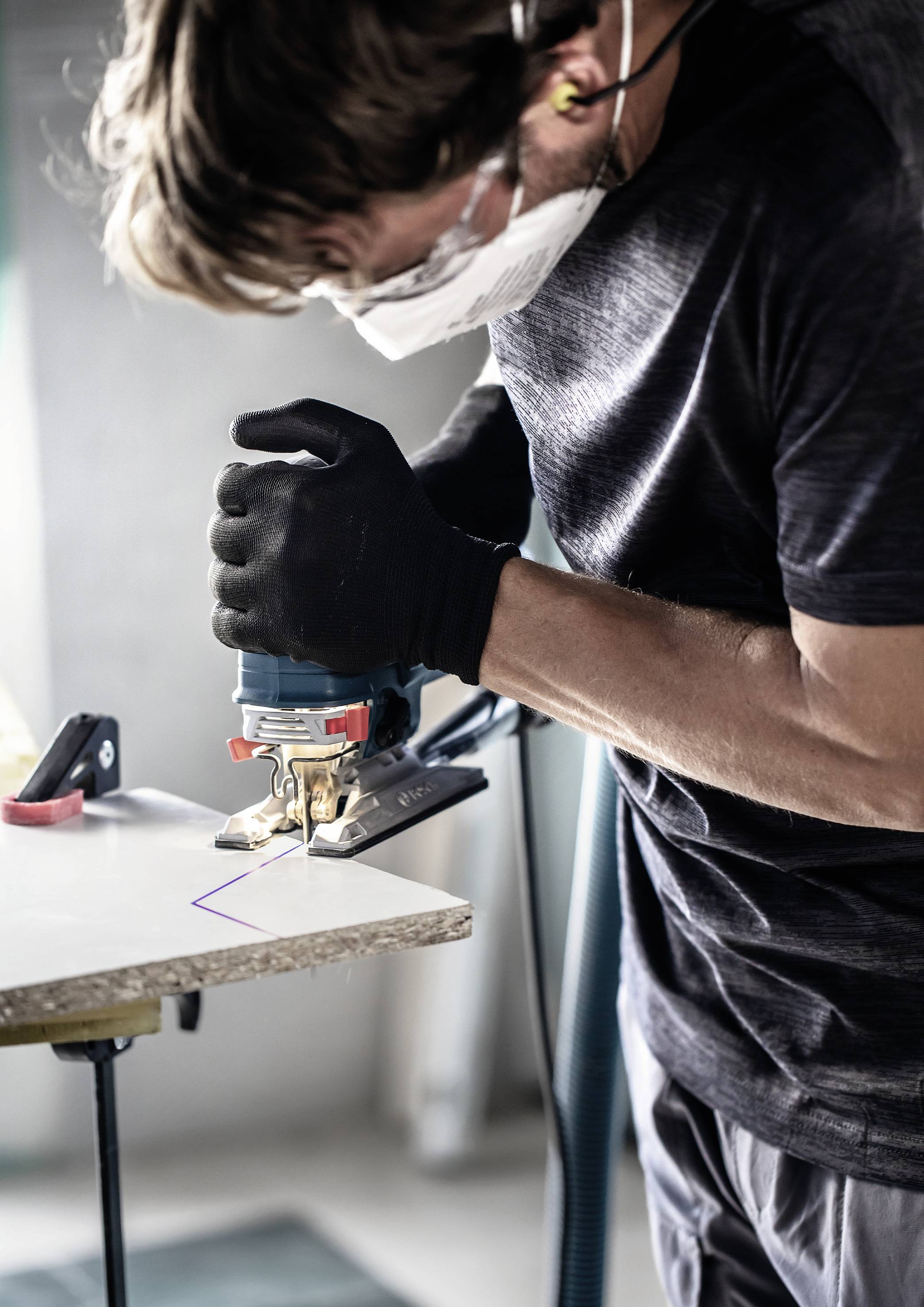 Eine Person trägt eine Schutzmaske und Handschuhe und verwendet eine elektrische Stichsäge, um Holz zu schneiden.