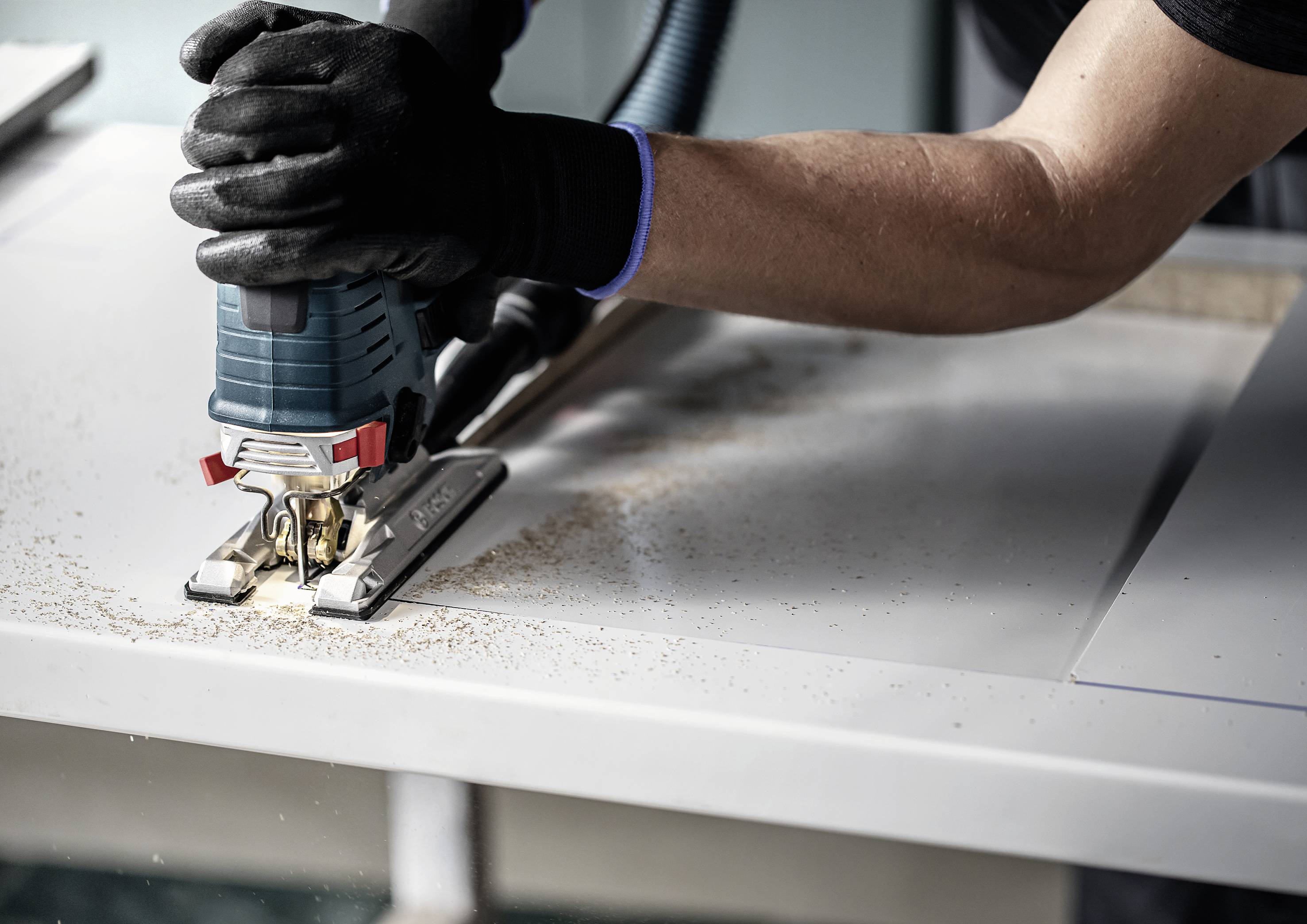Eine Person schneidet mit einer elektrischen Stichsäge in eine Holzplatte. Holzspäne fallen um das Schnittgebiet herum.