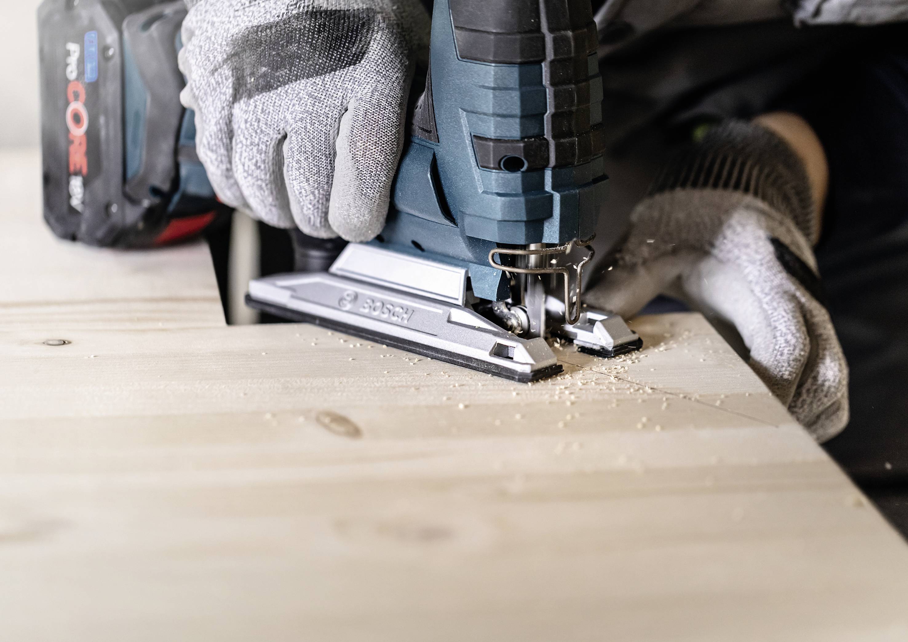 Eine Person sägt mit einer Stichsäge ein Holzbrett. Sie trägt Handschuhe zur Sicherheit. Holzspäne befinden sich nahe der Sägekante.