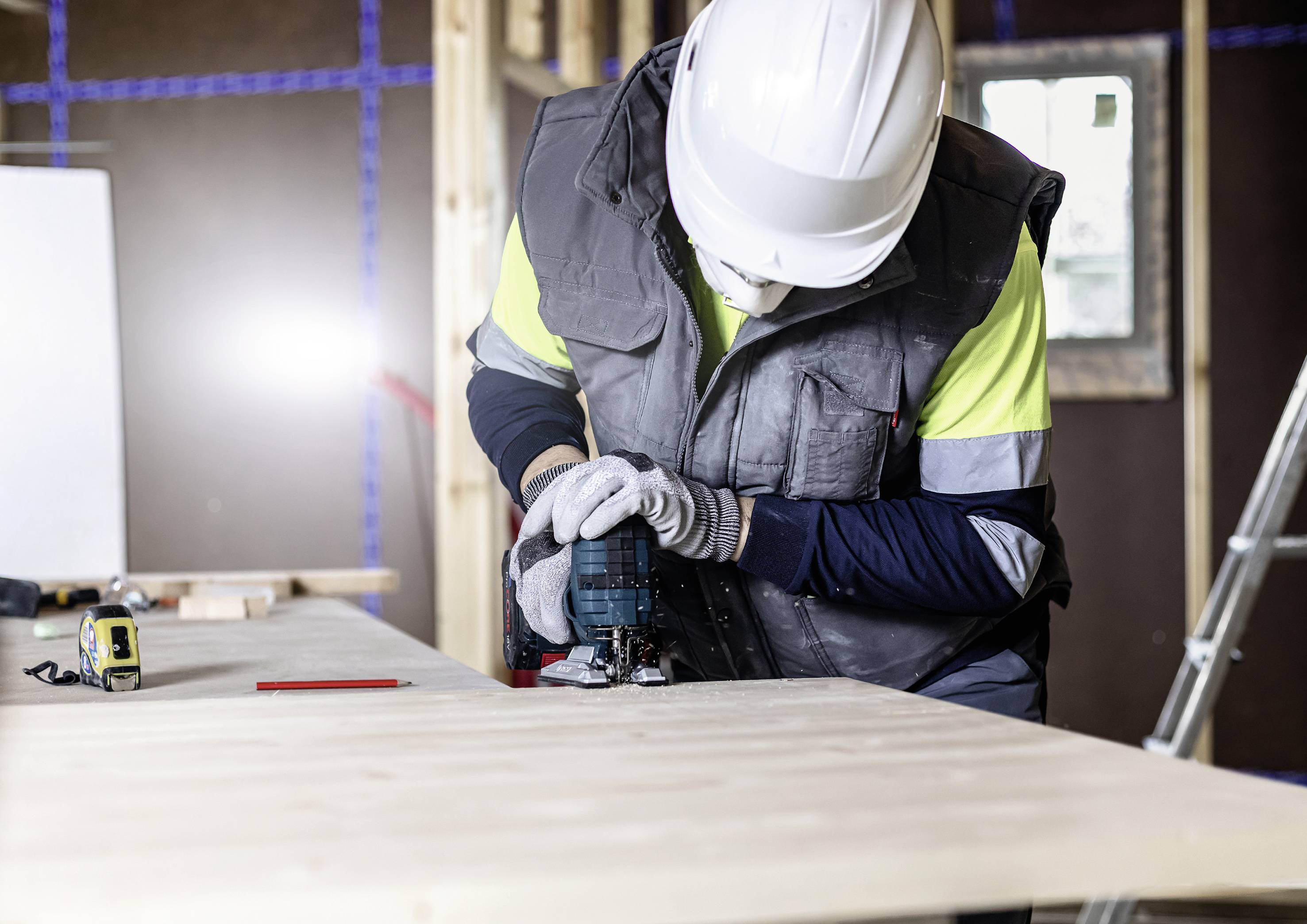 Ein Bauarbeiter mit Schutzhelm und Handschuhen bearbeitet Holz mit einer Elektrosäge in einer Baustellenumgebung.