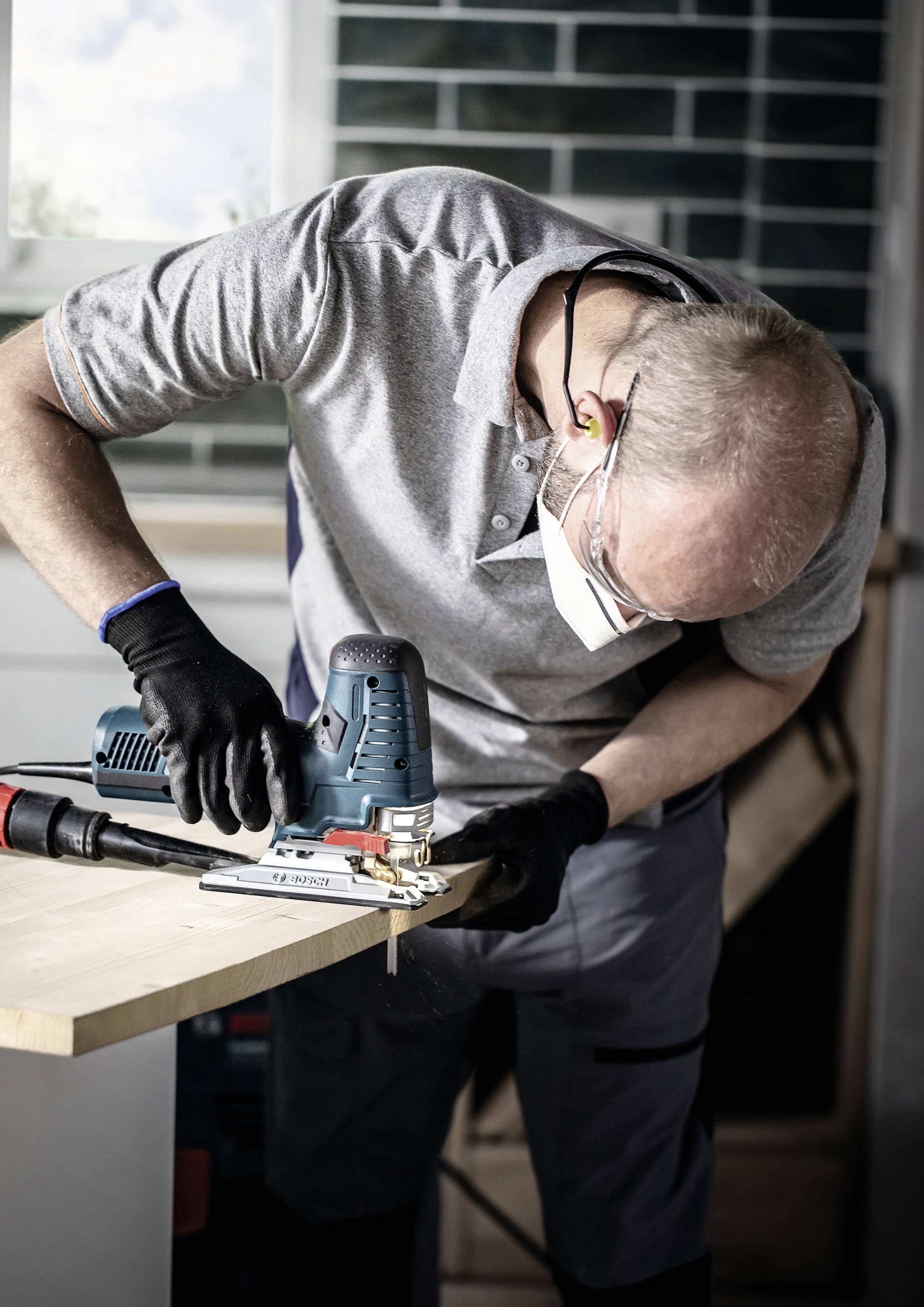 Ein Mann in Schutzbekleidung verwendet eine elektrische Säge, um ein Holzbrett zu schneiden, in einer Werkstattumgebung.