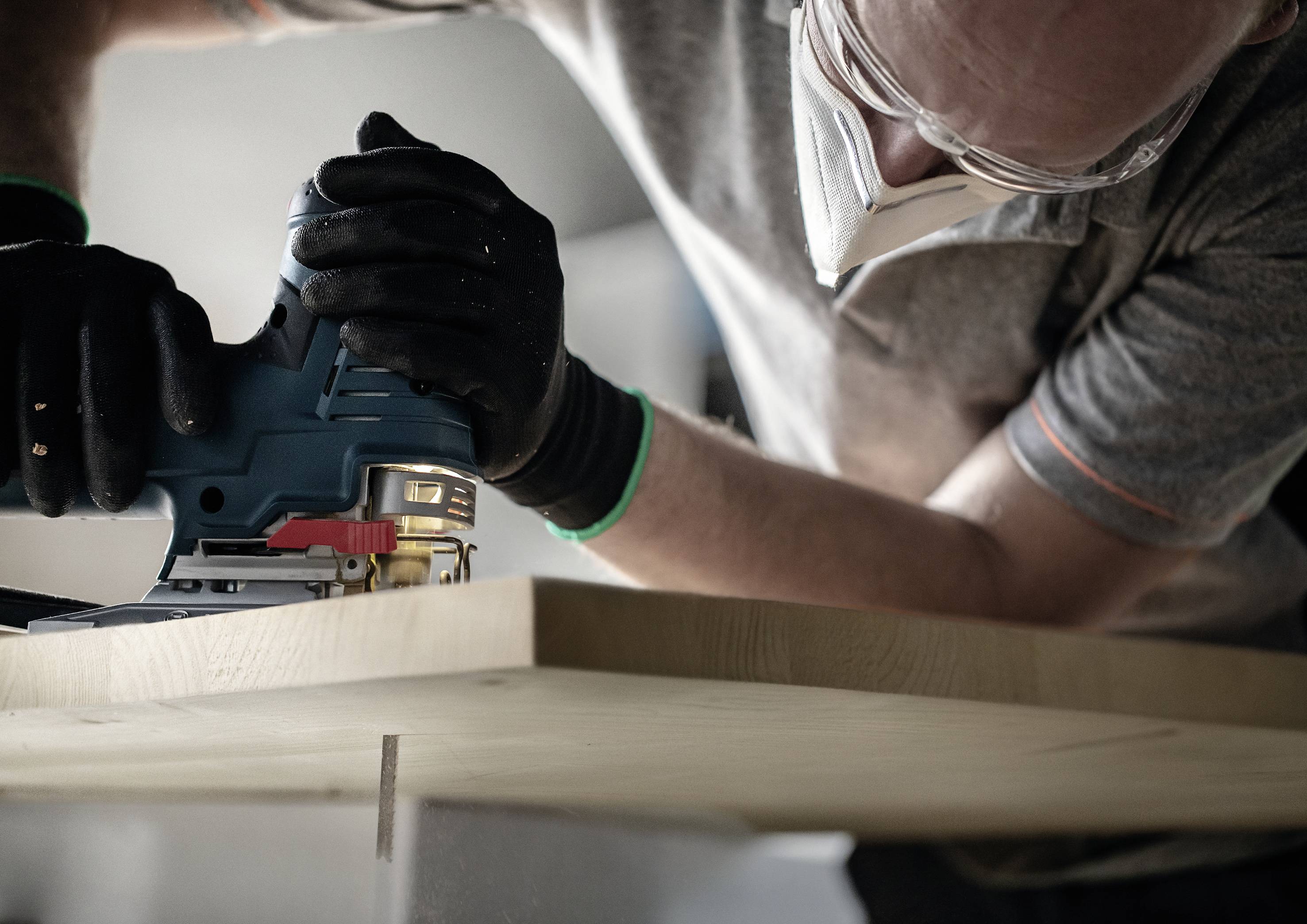 Eine Person trägt Schutzbrille und Handschuhe, während sie ein Stück Holz mit einer elektrischen Stichsäge schneidet.