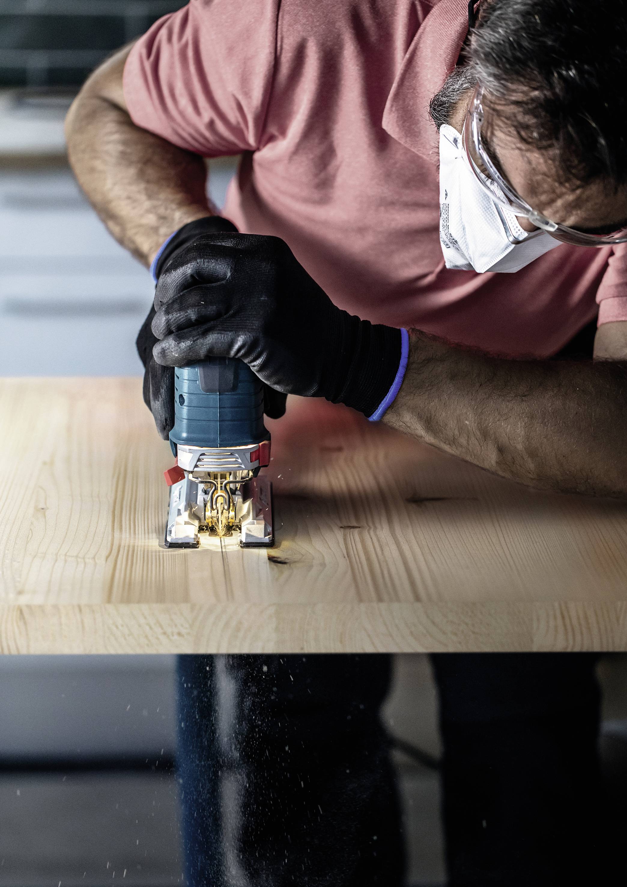 Ein Mann in Schutzmaske und Handschuhen benutzt eine elektrische Stichsäge, um ein Holzbrett zu schneiden.