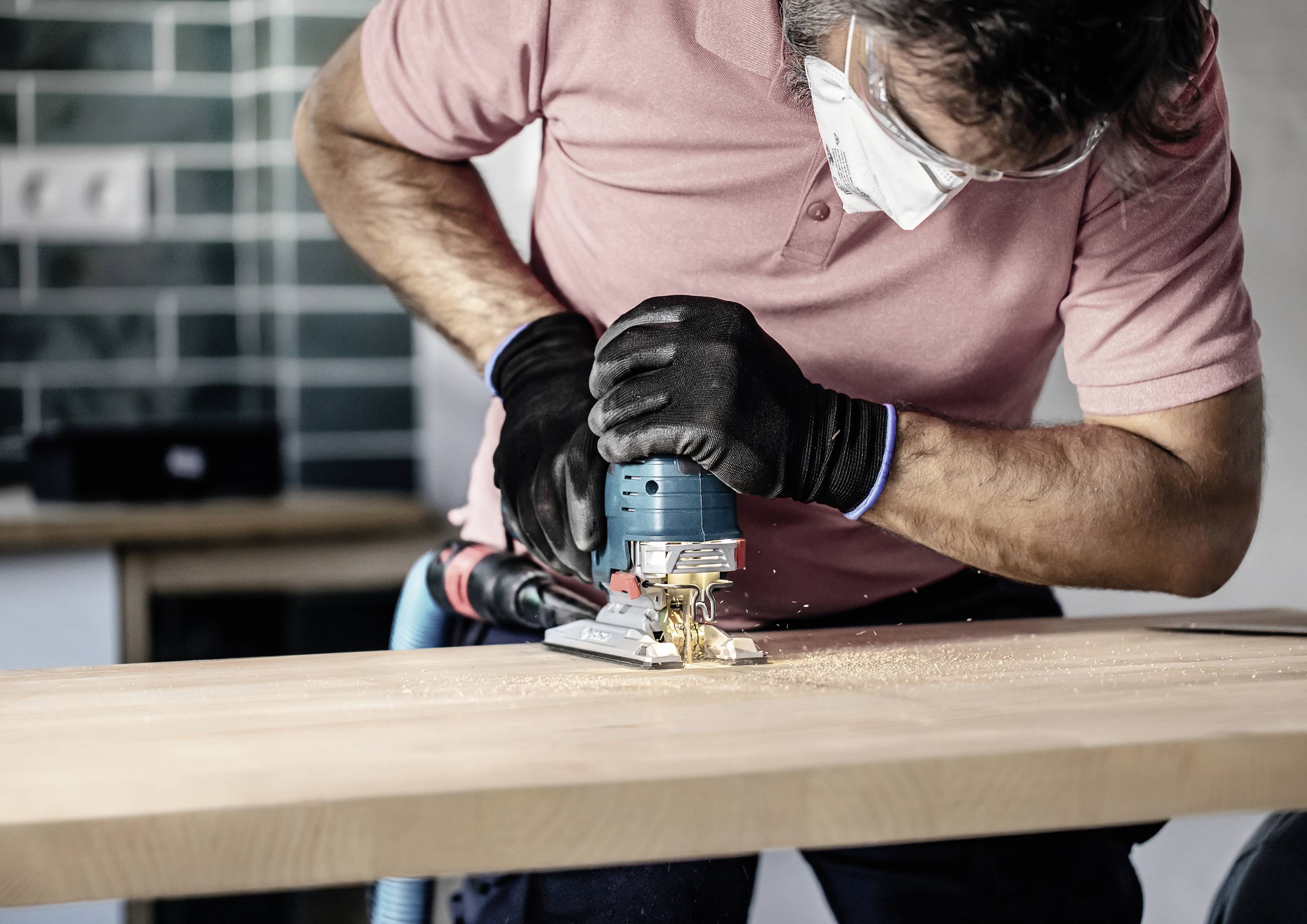Eine Person schleift mit einer Maschine eine Holzkante. Sie trägt Schutzbrille und Maske. Funken sprühen vom Holz, das sich auf einem Tisch befindet.