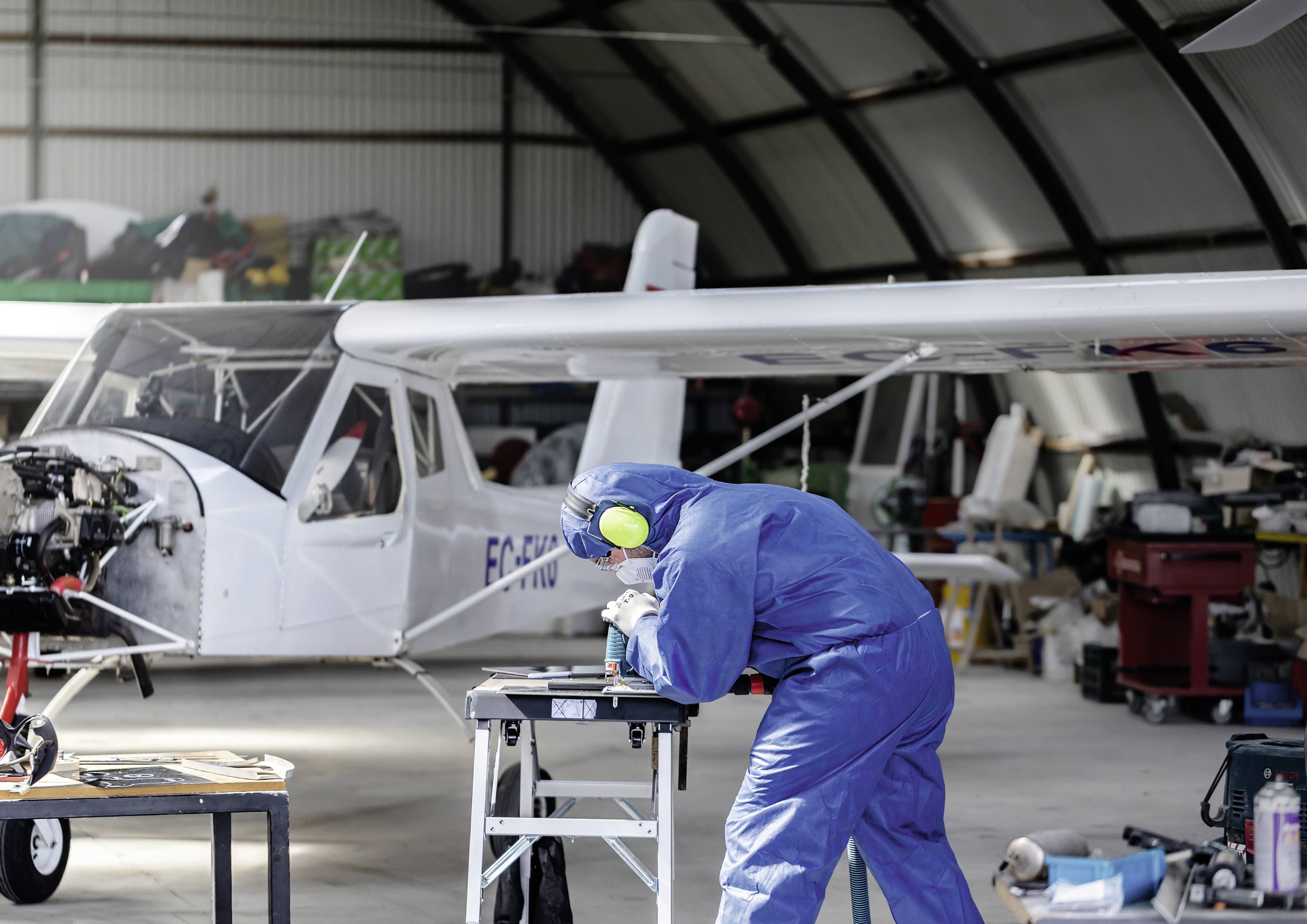 Ein Techniker im blauen Schutzanzug arbeitet an einem Arbeitsplatz in einem Hangar. Ein kleines Flugzeug ist im Hintergrund sichtbar.