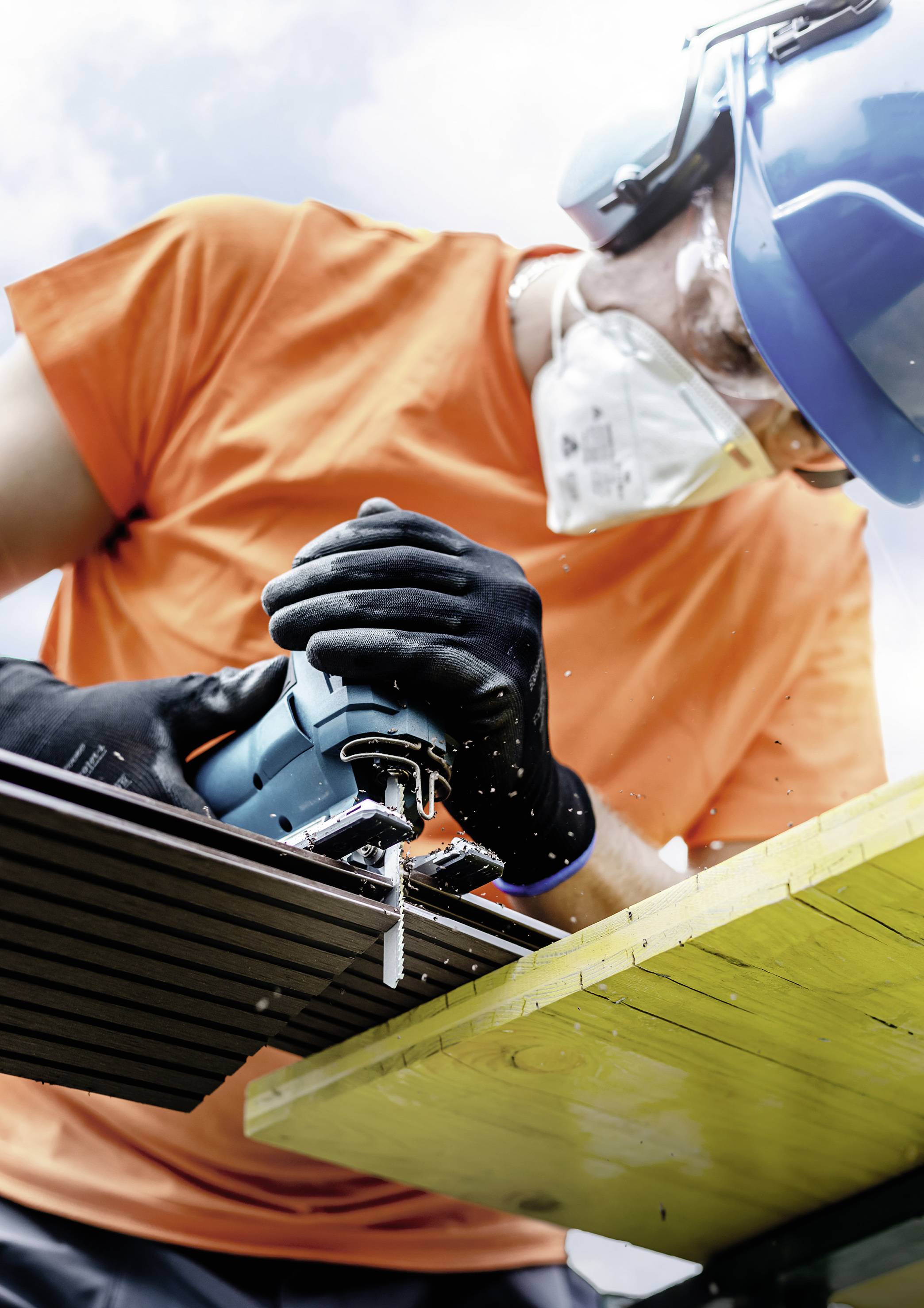 Eine Person mit Schutzkleidung, Helm und Maske bearbeitet Holz mit einer elektrischen Säge im Freien.
