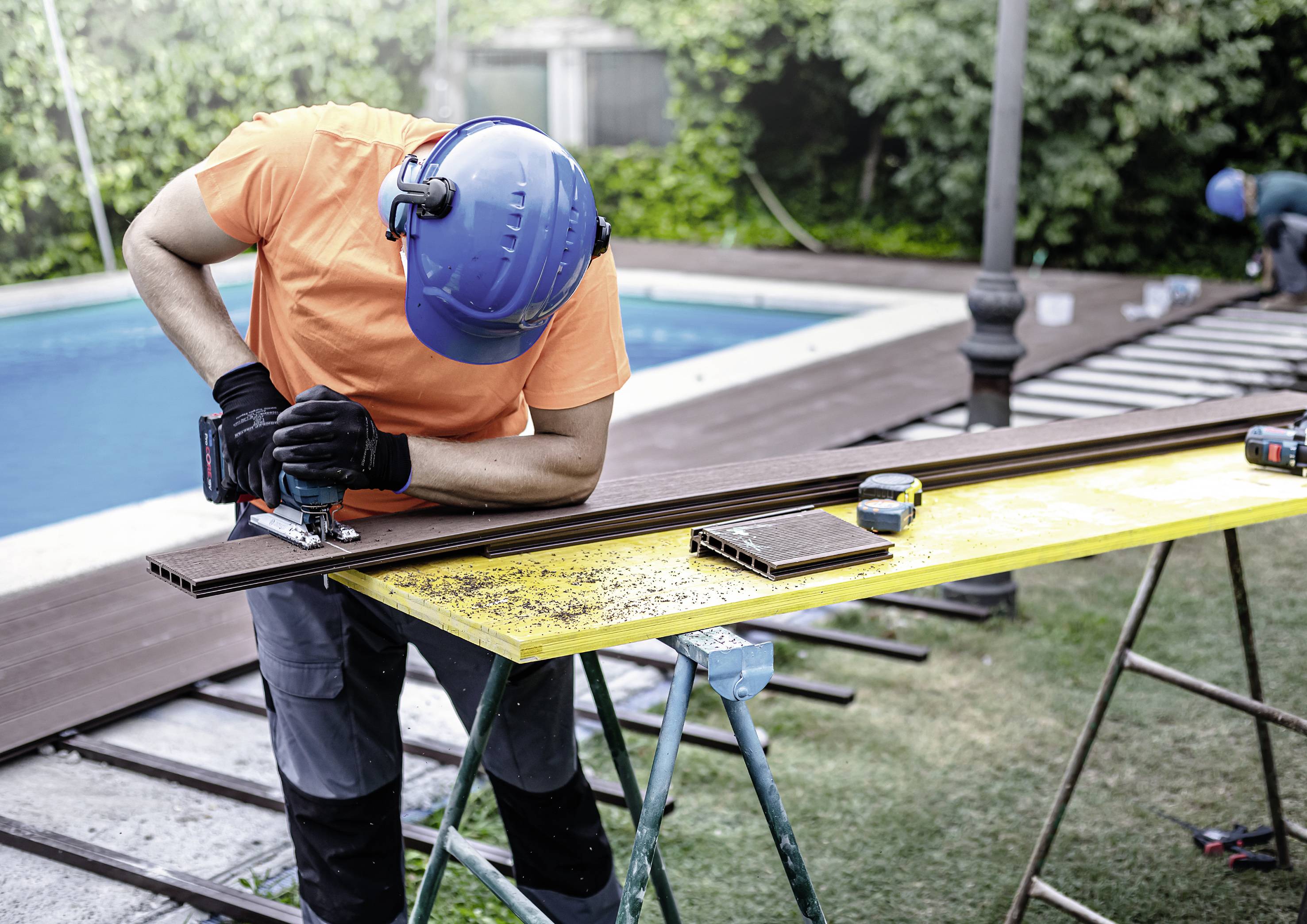 Arbeiter mit Helm und Gehörschutz sägt Terrassendielen neben einem Pool auf einem Tisch im Freien.