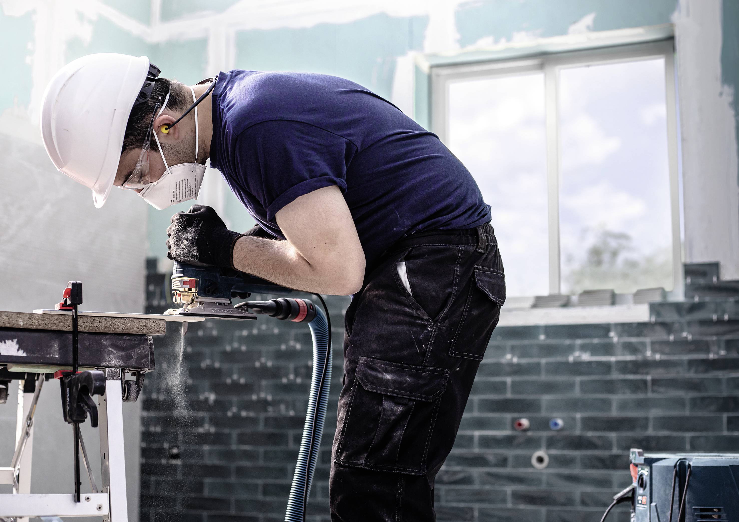 Ein Arbeiter mit Helm und Maske benutzt ein Elektrowerkzeug, um Holz in einem halb renovierten Raum zu schneiden.