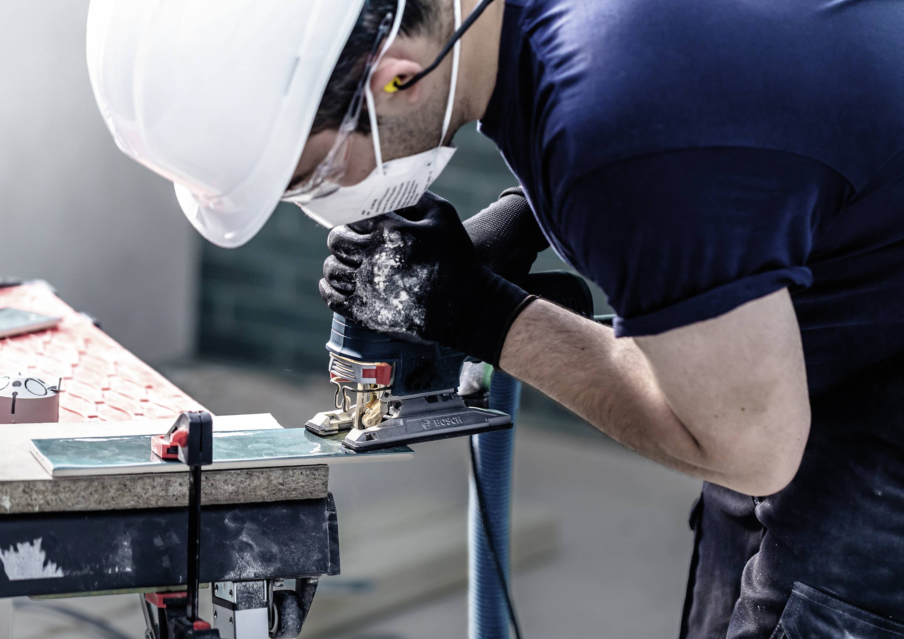 Eine Person mit Schutzhelm und Maske arbeitet mit einem Elektrowerkzeug an einem Holzstück in einer Werkstattumgebung.