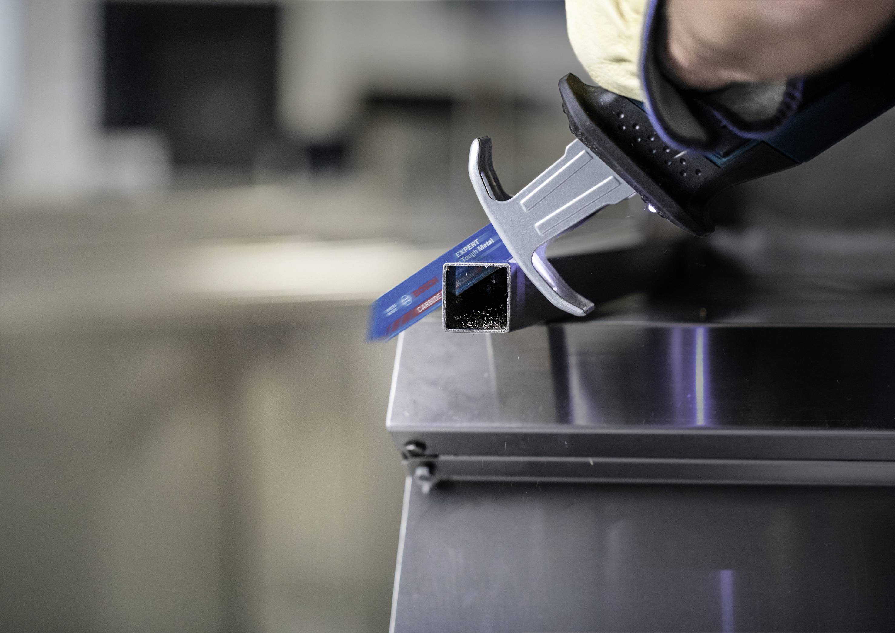 Eine Hand in einem Handschuh hält eine digitale Schieblehre, die die Breite eines Metallrohrs auf einer Werkbank misst.