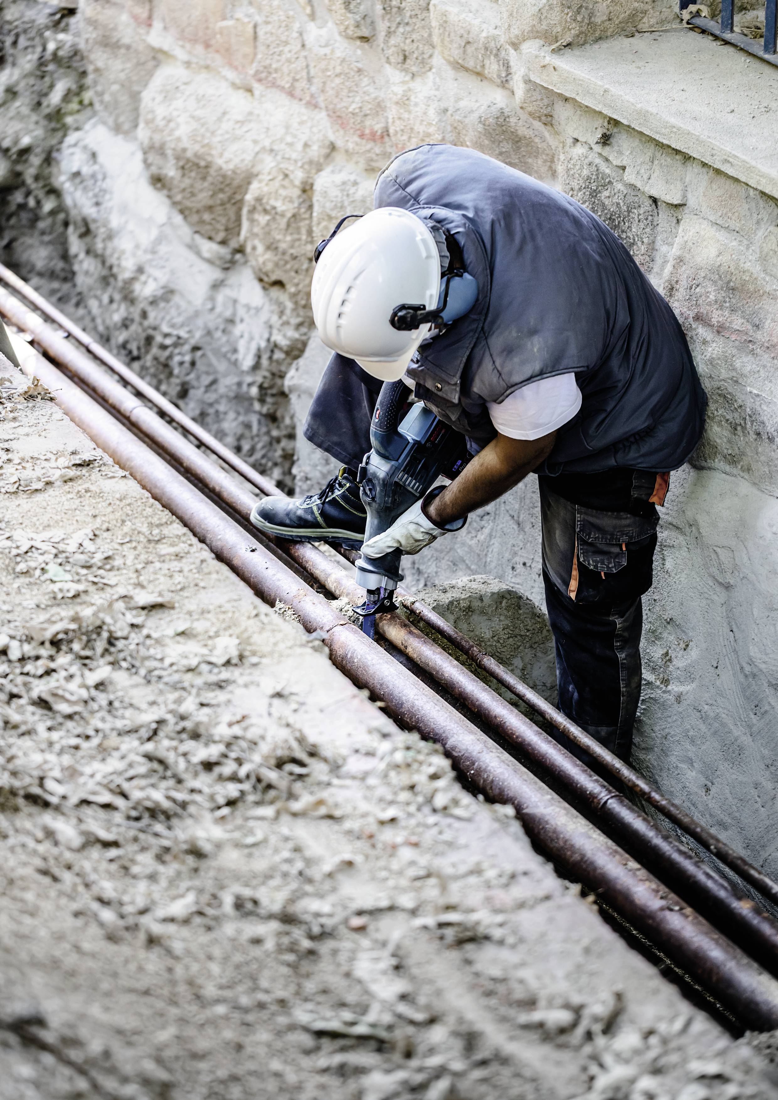 Ein Arbeiter mit Schutzhelm und Handschuhen repariert Rohrleitungen mit einem Bohrwerkzeug in einem Graben neben einer Mauer.
