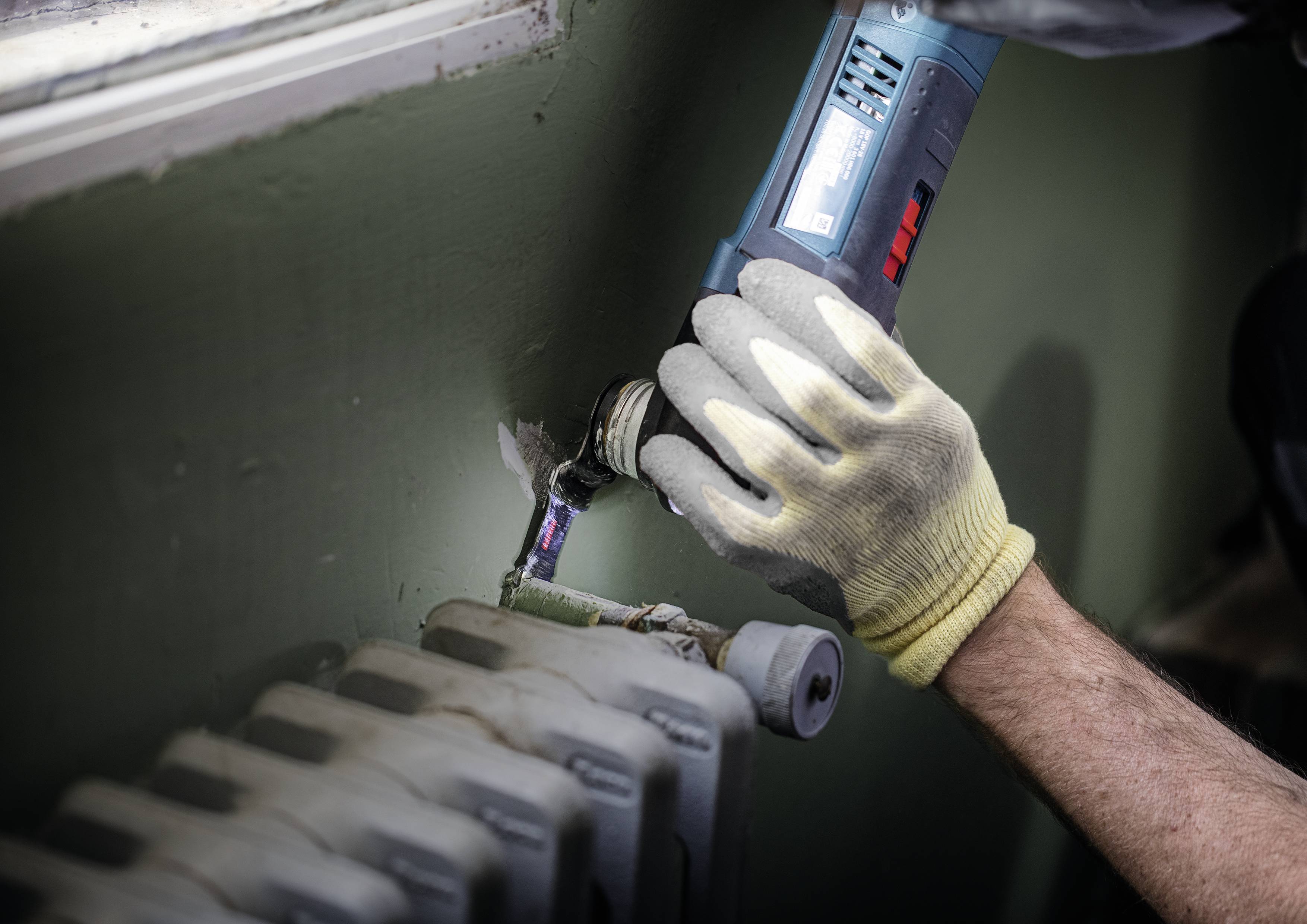 Eine Person repariert mit Handschuhen eine Heizungsleitung an der Wand mit einem blauen Werkzeug.