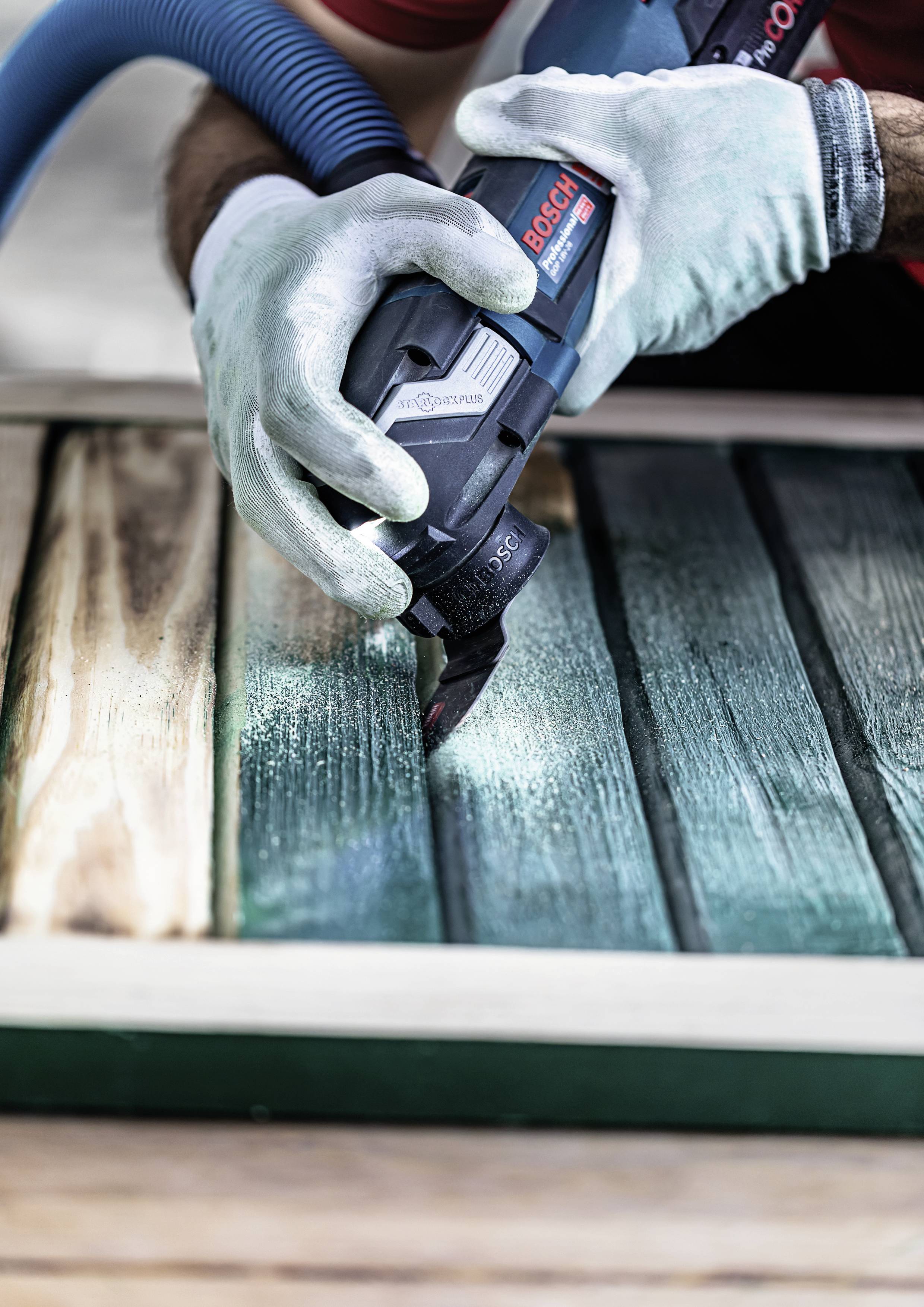 Person schleift mit Elektrowerkzeug Holzplanken. Blaue Schutzbrille und Handschuhe werden getragen; grünliches Holz wird bearbeitet.