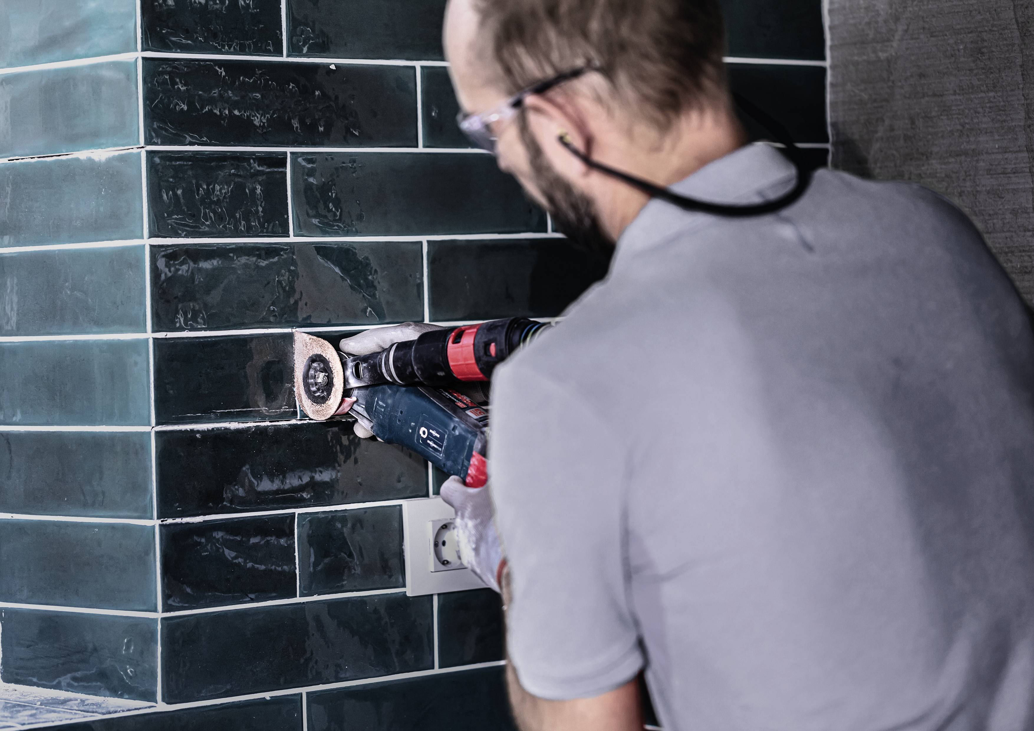 Ein Handwerker bohrt mit einer Maschine in eine geflieste Wand. Er trägt Schutzbrille und Gehörschutz. Die Fliesen sind dunkelgrün.