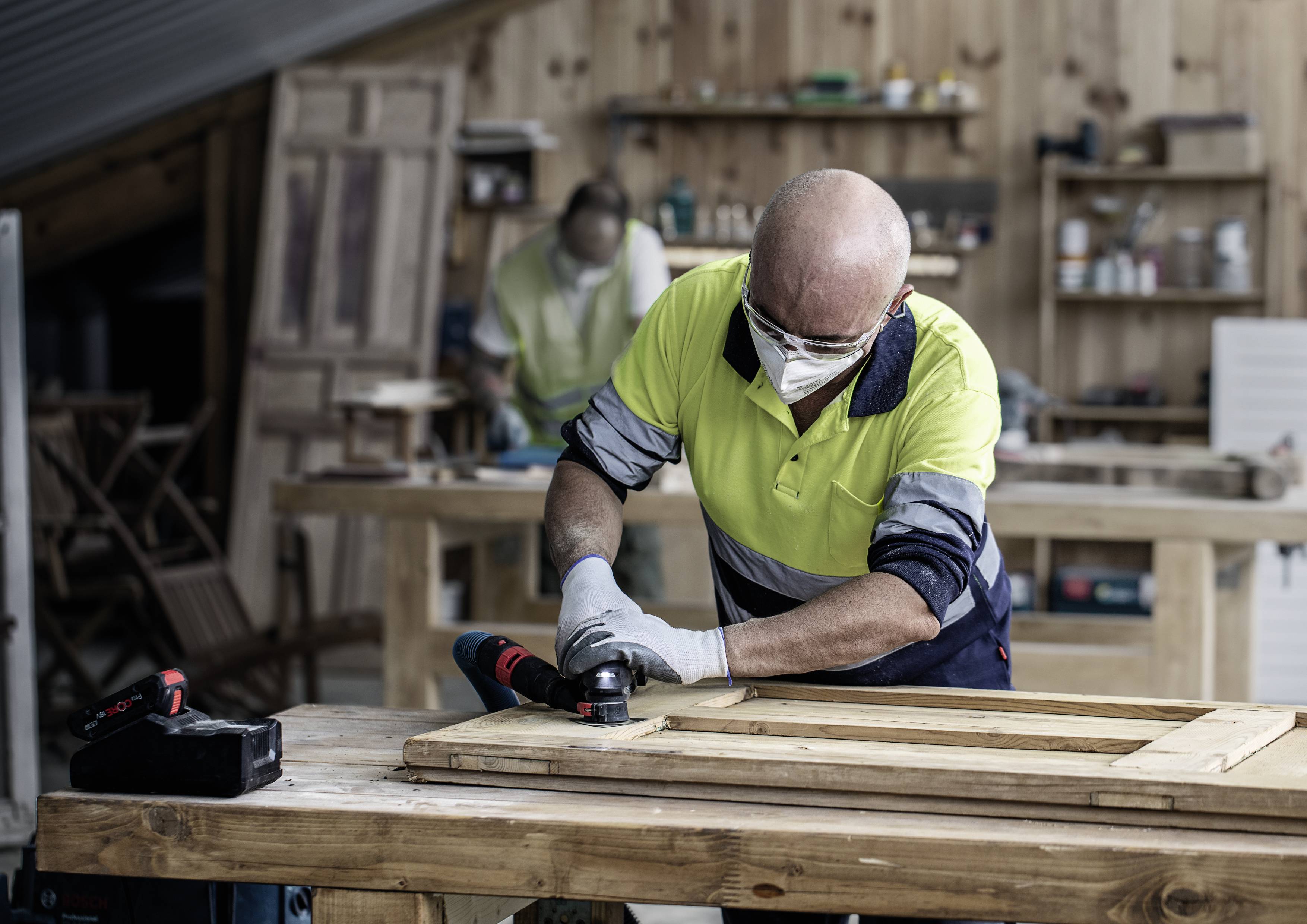 Ein Mann in Arbeitskleidung und Schutzmaske arbeitet an einem Holzstück in einer Werkstatt. Im Hintergrund steht eine weitere Person.