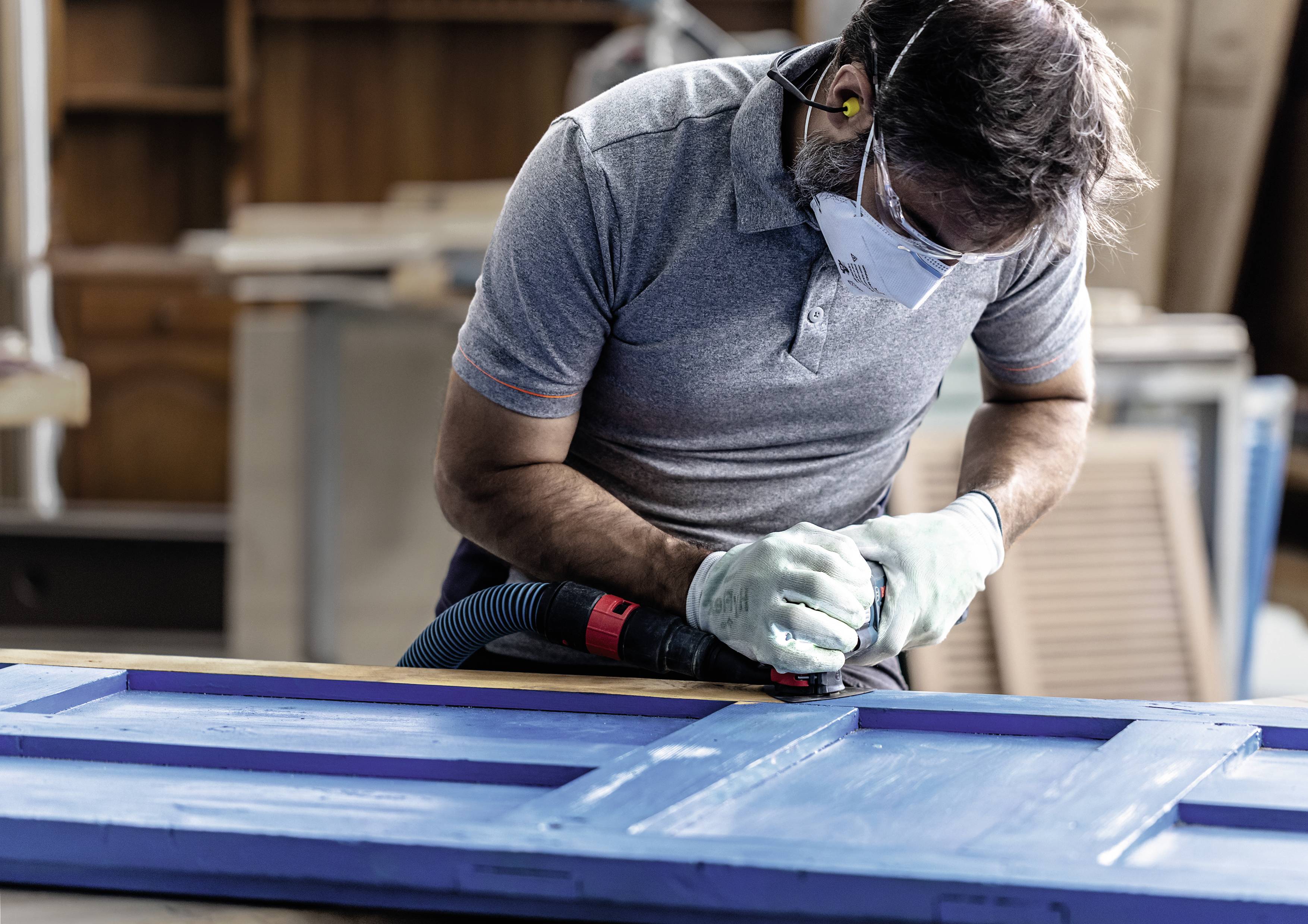 Ein Mann arbeitet konzentriert mit Schutzbrille und Maske an einer blauen Holztür, die er mit einem Schleifgerät bearbeitet.