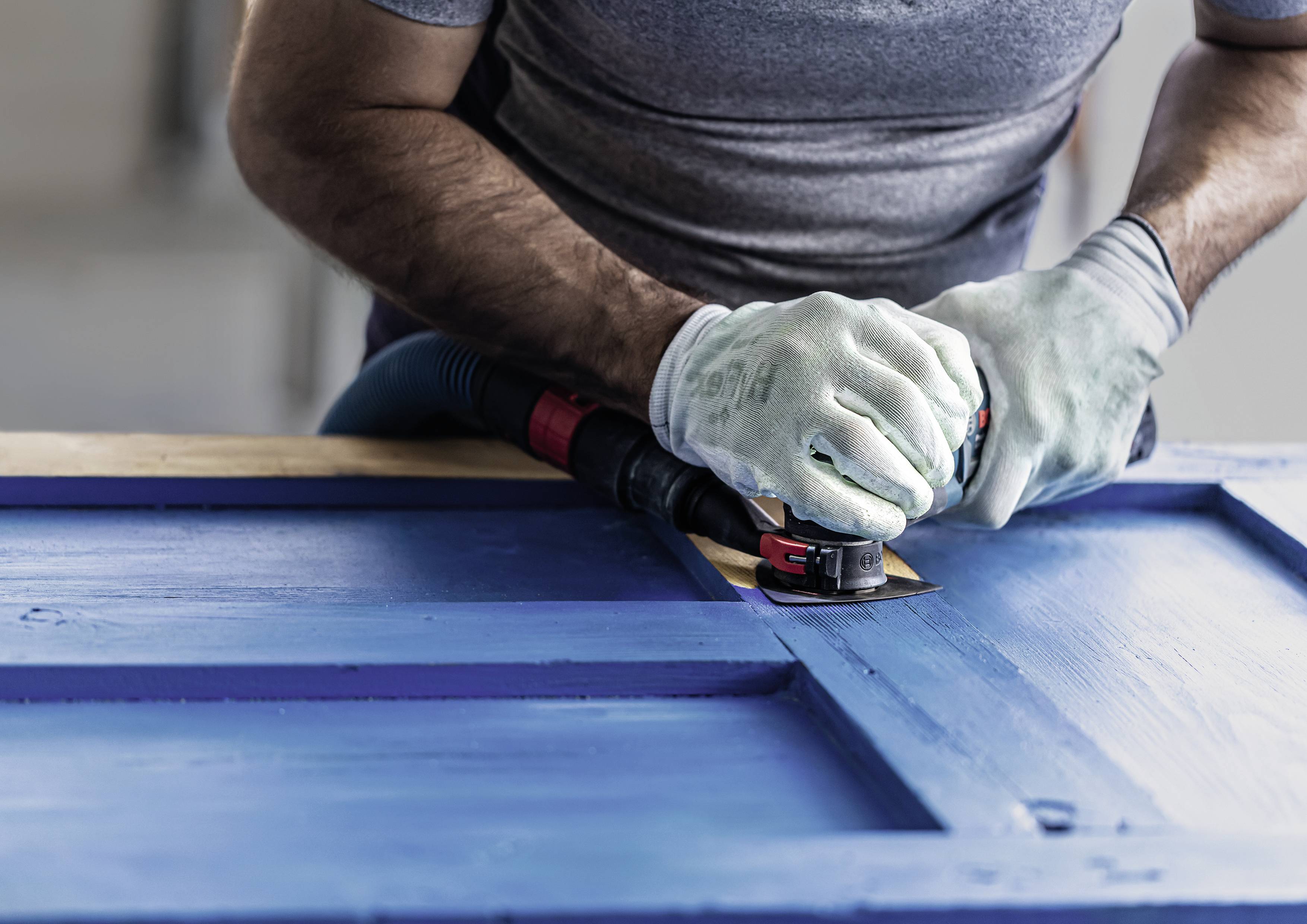 Eine Person schleift mit einem Handschleifer blaues Holz, möglicherweise eine Tür. Sie trägt Schutzhandschuhe.