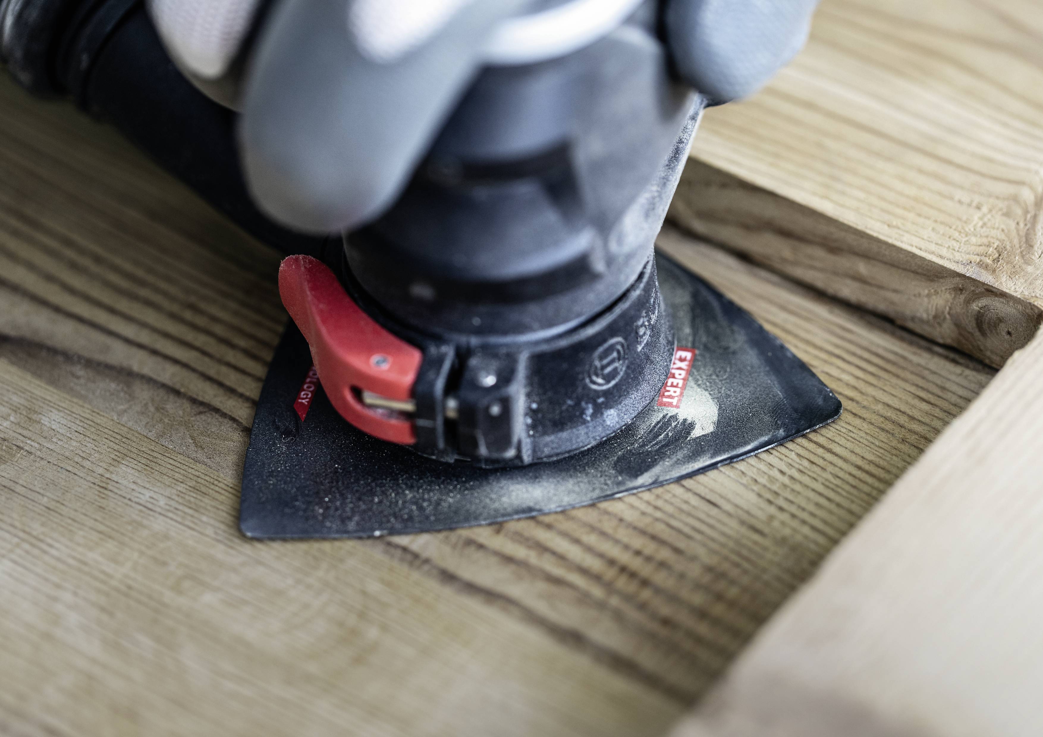 Eine Hand mit Handschuh hält ein elektrisches Schleifgerät, das Holzoberfläche abschleift. Gerät hat eine dreieckige schwarze Schleiffläche.
