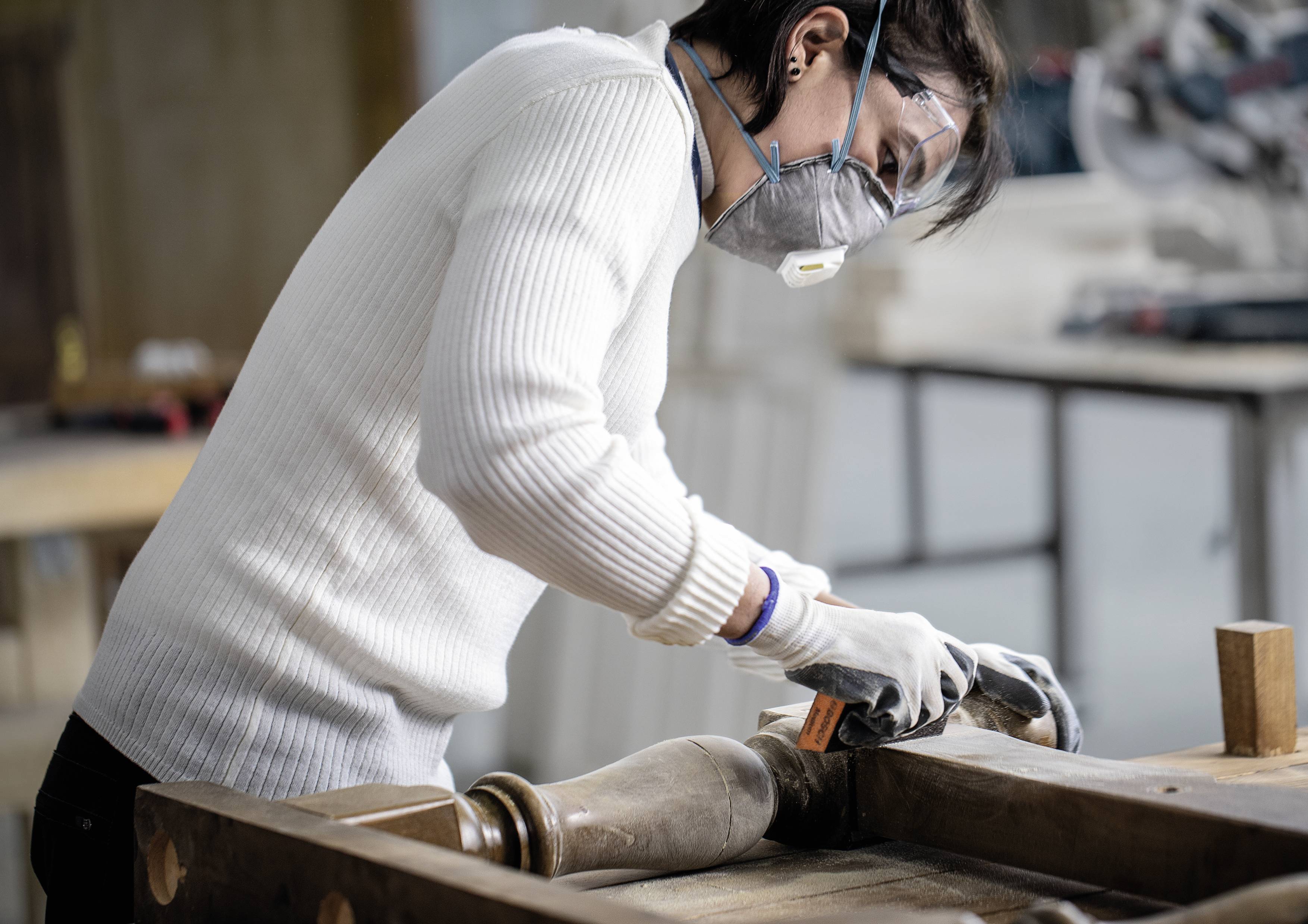 Eine Person in Schutzkleidung bearbeitet ein Holzstück in einer Werkstatt. Sie trägt Maske und Brille zum Schutz.