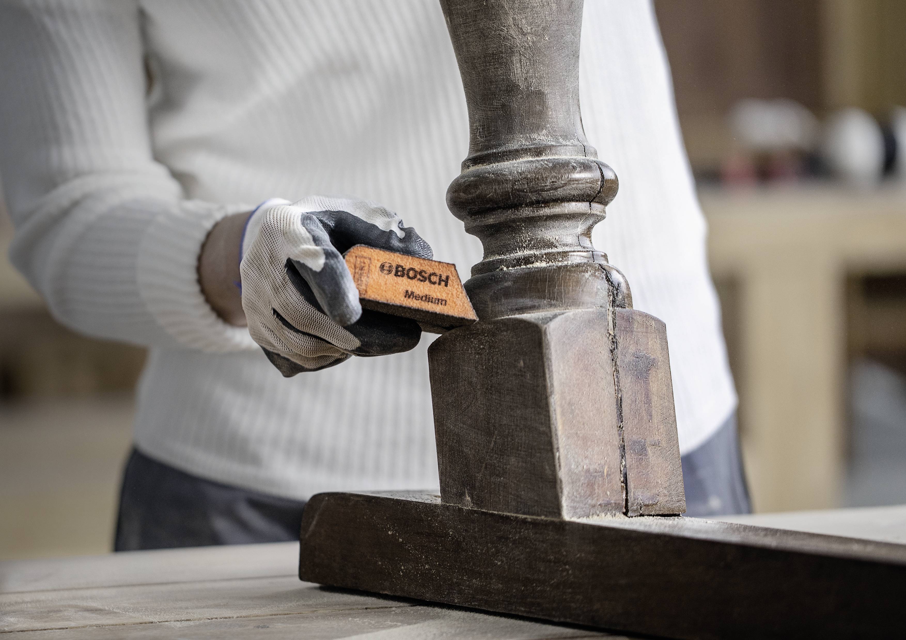 Eine Person schleift mit einem 'BOSCH'-Schleifblock eine Holzsäule. Die Person trägt einen weißen Pullover und Arbeitshandschuhe.