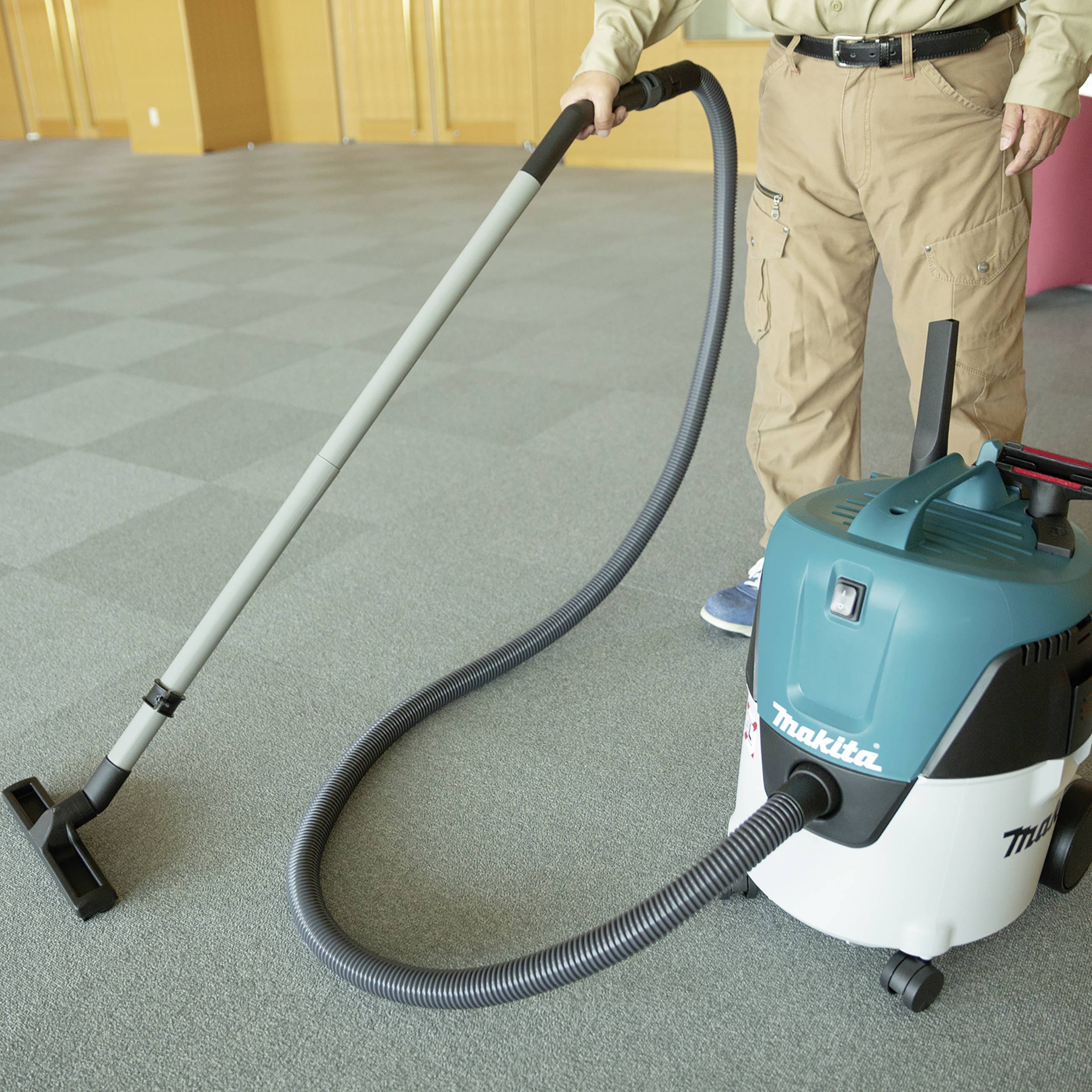Person mit khakifarbenen Hosen benutzt einen Staubsauger auf einem Teppichboden in einem Büro.