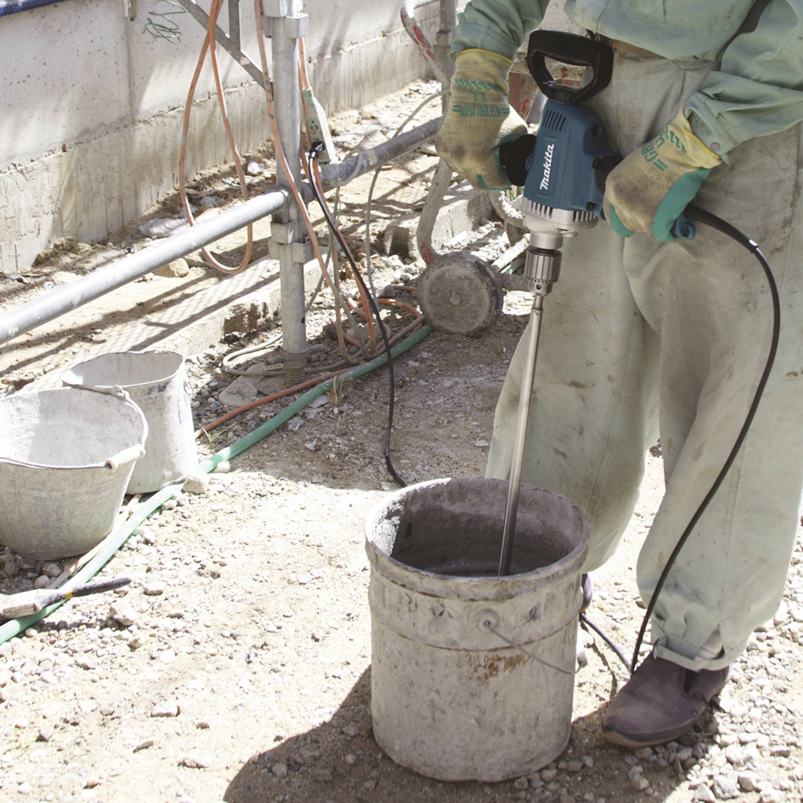 Person mischt Zement mit einer elektrischen Mischmaschine in einem Eimer auf einer Baustelle. Schutzkleidung und Handschuhe werden getragen.