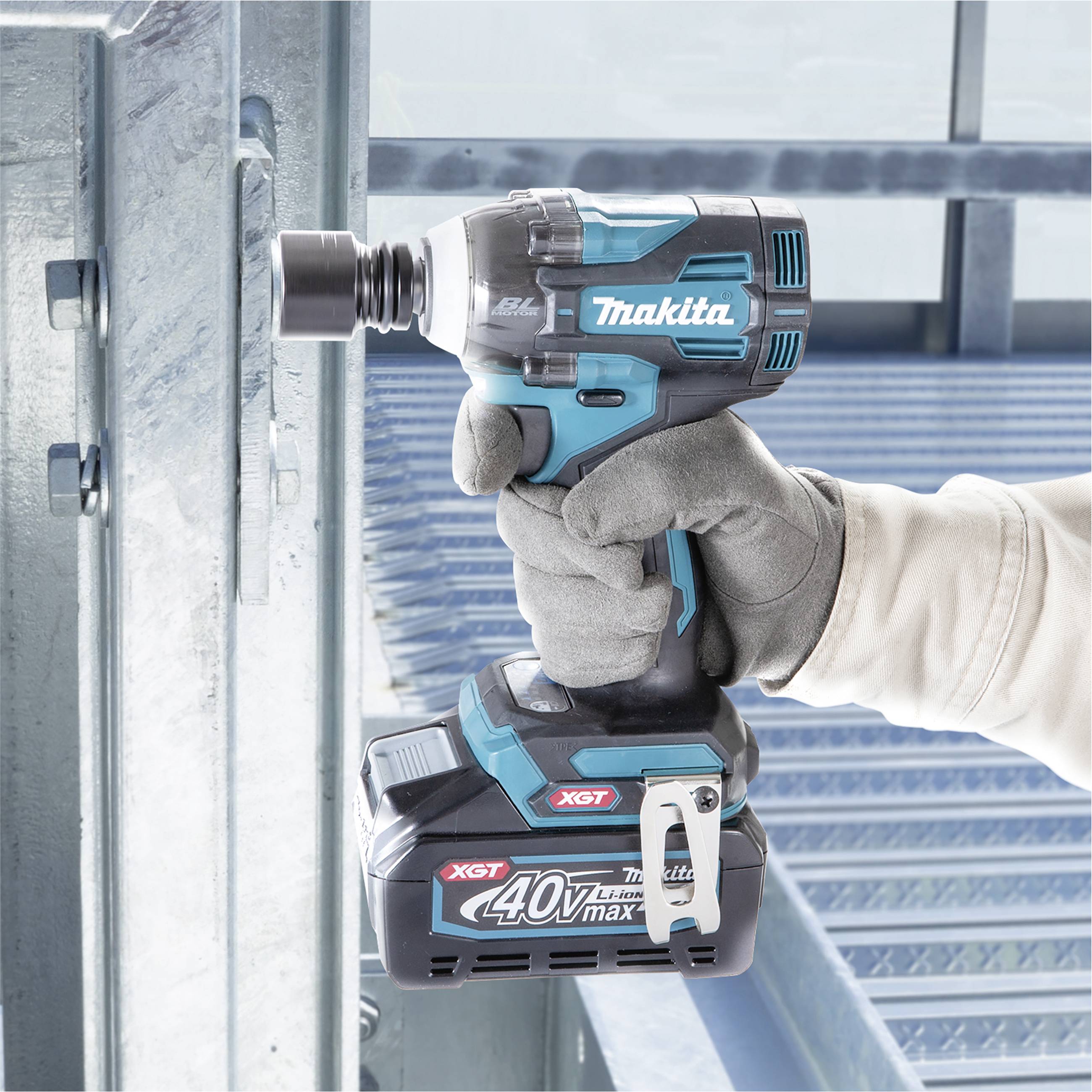 Eine Person mit Arbeitshandschuh hält einen blauen Akkuschrauber, der auf eine Metallkonstruktion zielt, mit 'Makita' Logo sichtbar.
