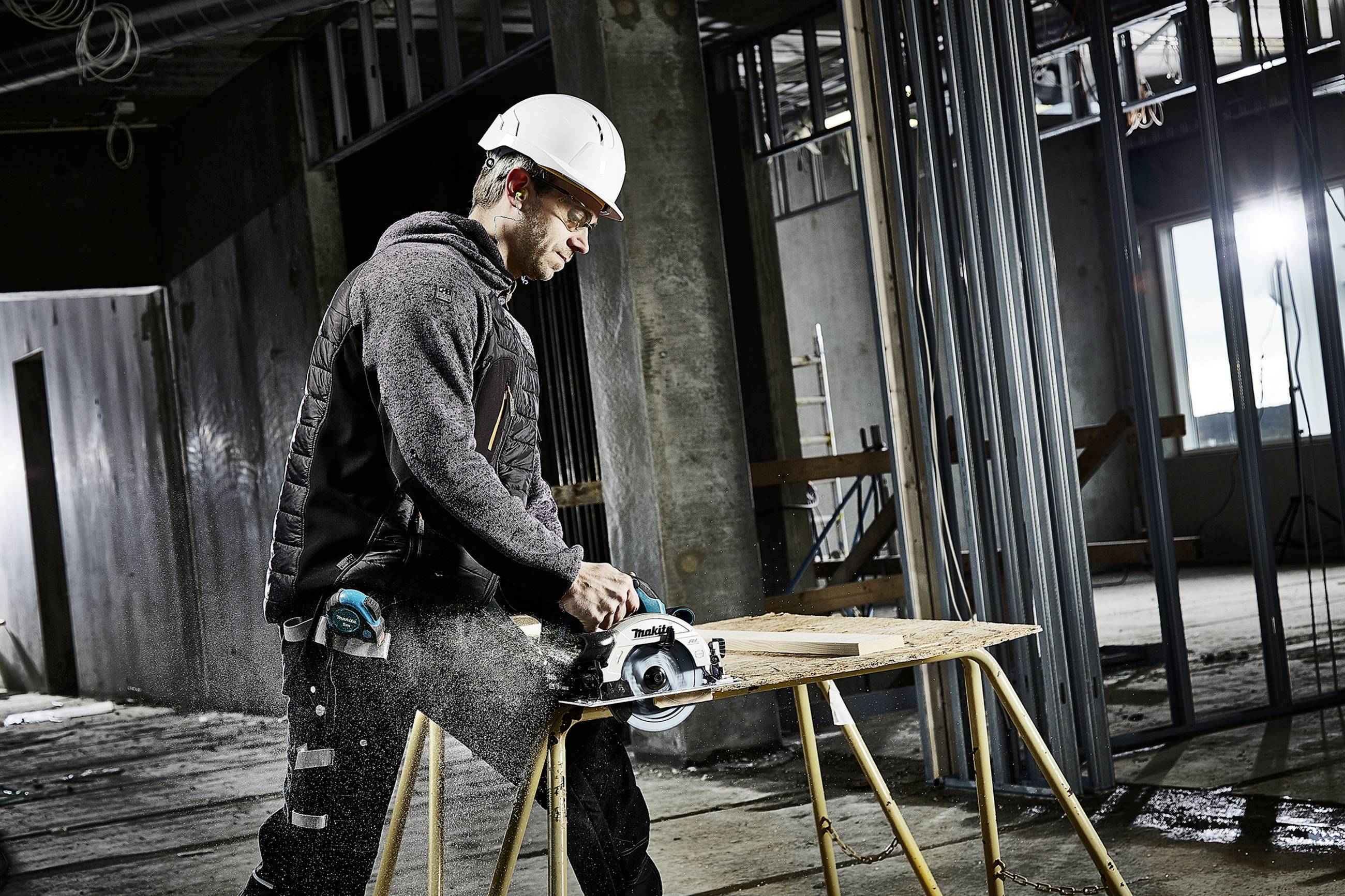Ein Bauarbeiter mit Schutzhelm schneidet Holz mit einer Kreissäge in einem unfertigen Gebäuderaum. Baustelle im Hintergrund.
