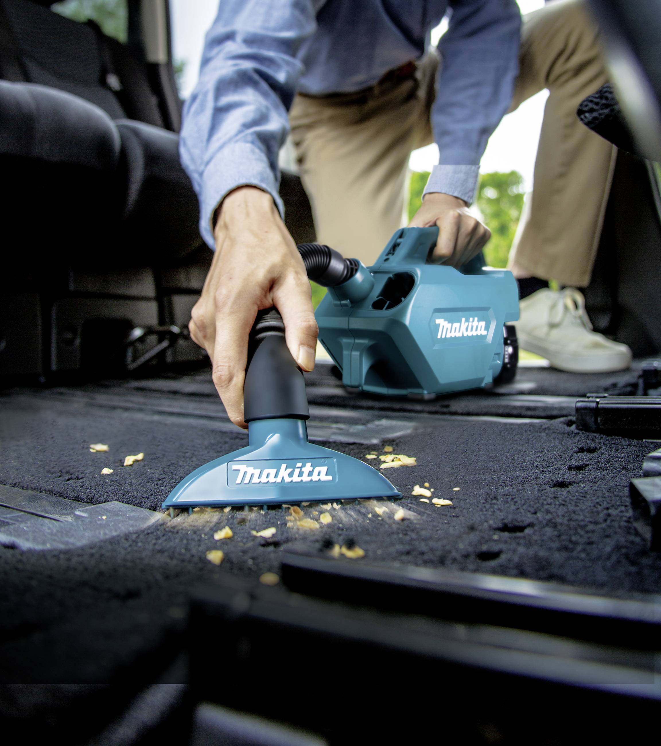 Eine Person benutzt einen blauen Handstaubsauger von Makita, um Krümel von einem schwarzen Teppich in einem Auto zu entfernen.
