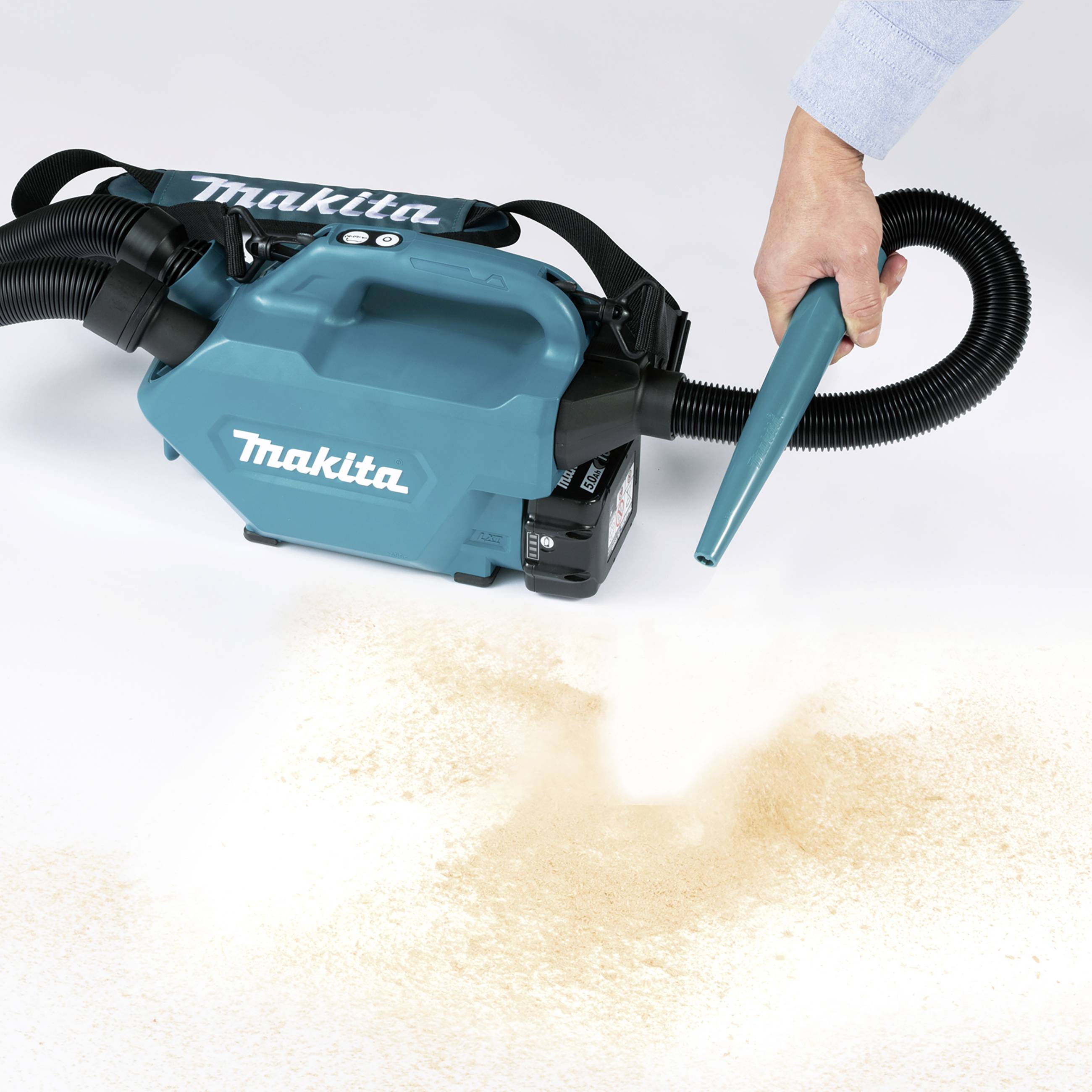 Ein Handstaubsauger von Makita saugt Holzspäne auf. Eine Person führt den Schlauch über eine Oberfläche mit verstreuten Spänen.