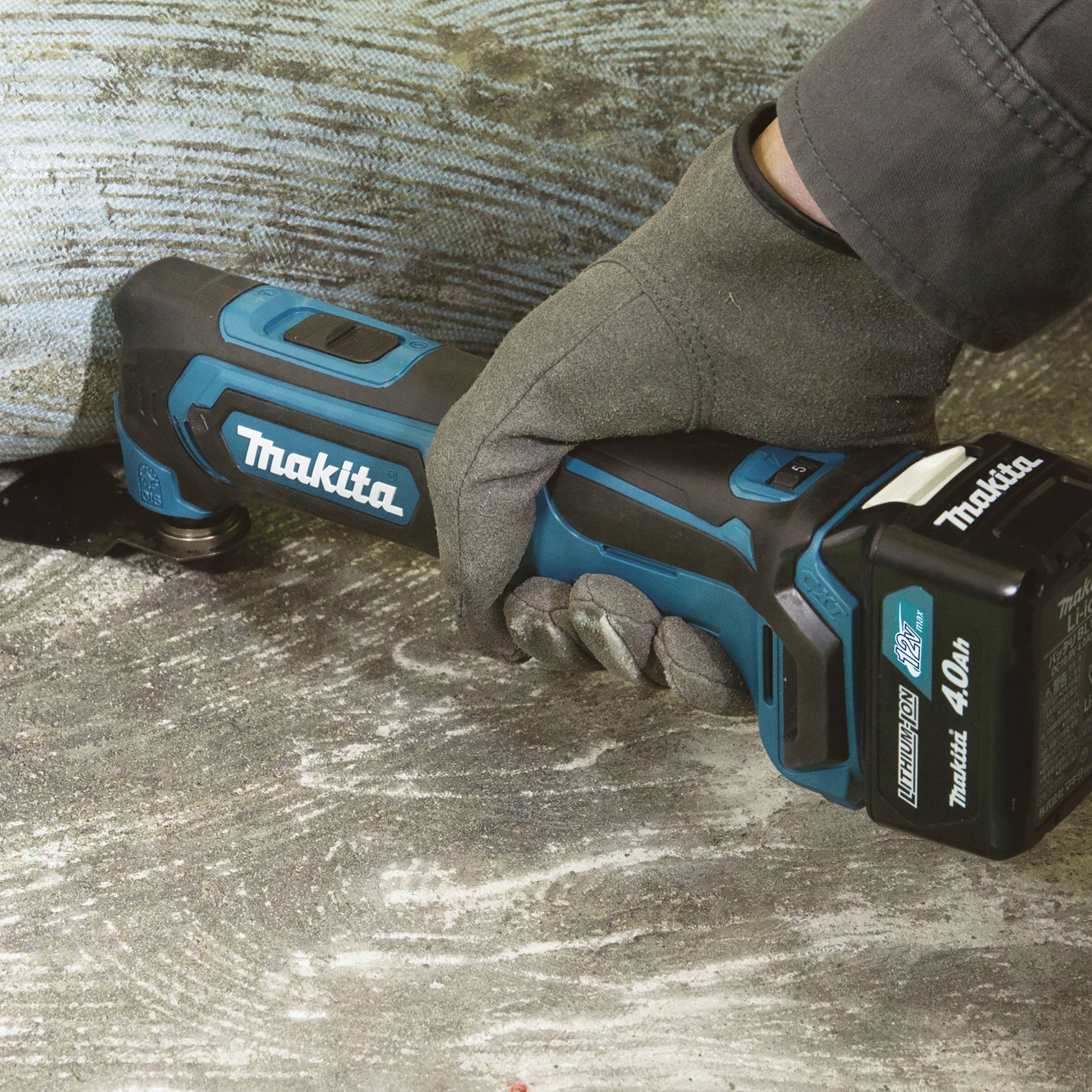 Eine Person hält ein elektrisches Handwerkzeug von Makita mit blau-schwarzem Gehäuse und Akku, beim Arbeiten auf einem Bodenbelag.