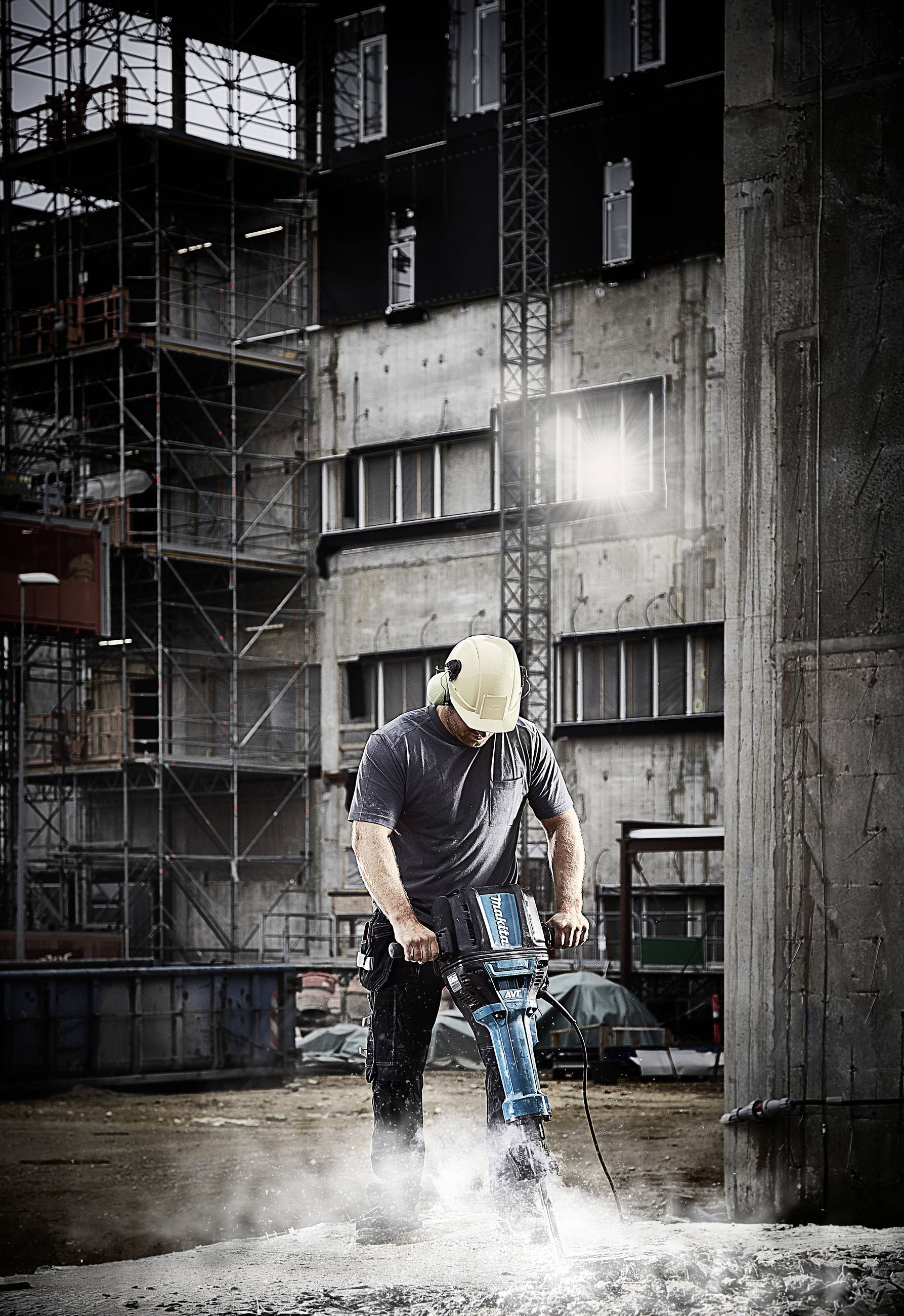 Eine Person arbeitet auf einer Baustelle mit einem Presslufthammer. Im Hintergrund sind Gerüste und Gebäude sichtbar.