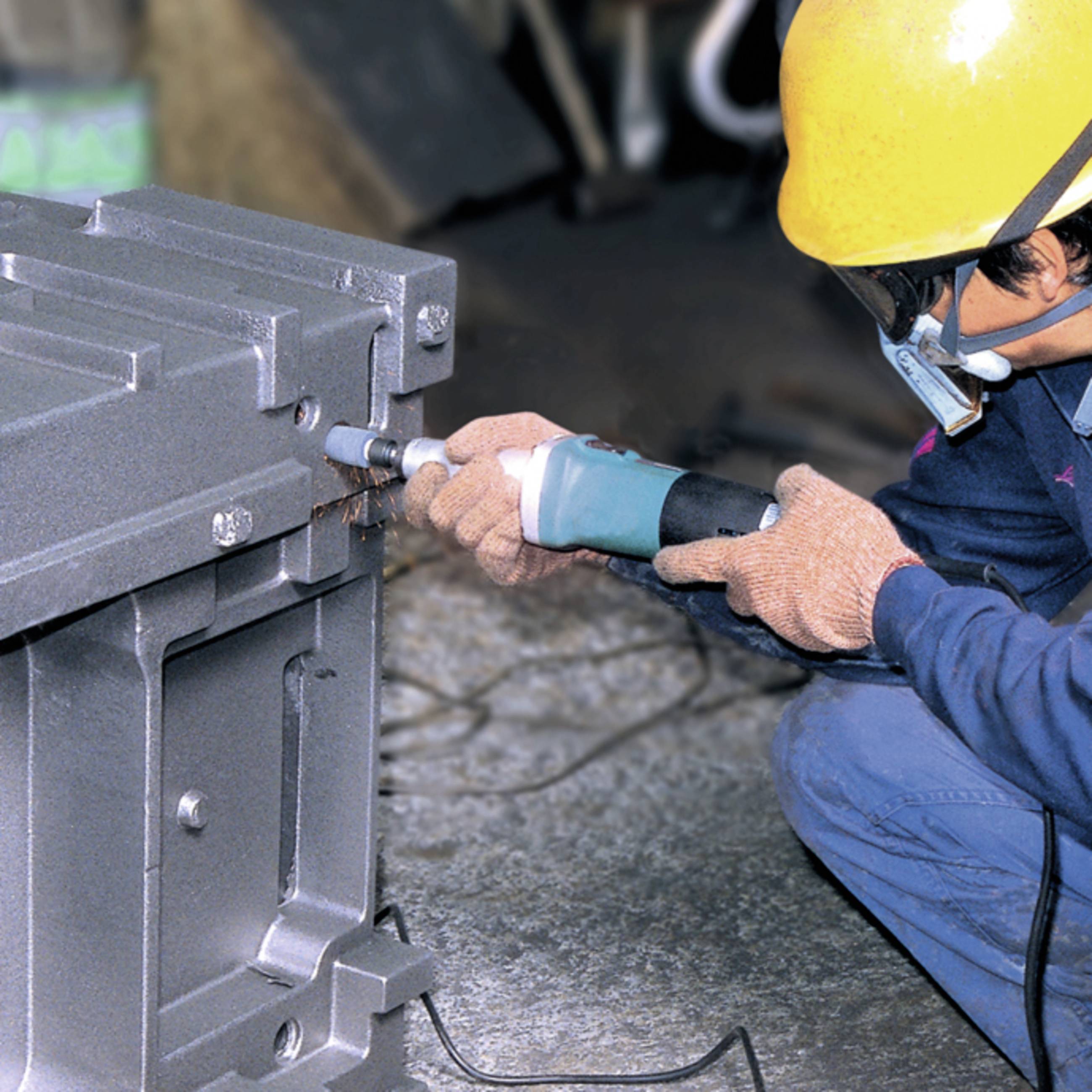 Eine Person mit Schutzhelm arbeitet mit einem Bohrer an einem großen Metallblock, möglicherweise in einer Werkstatt oder Fabrikumgebung.