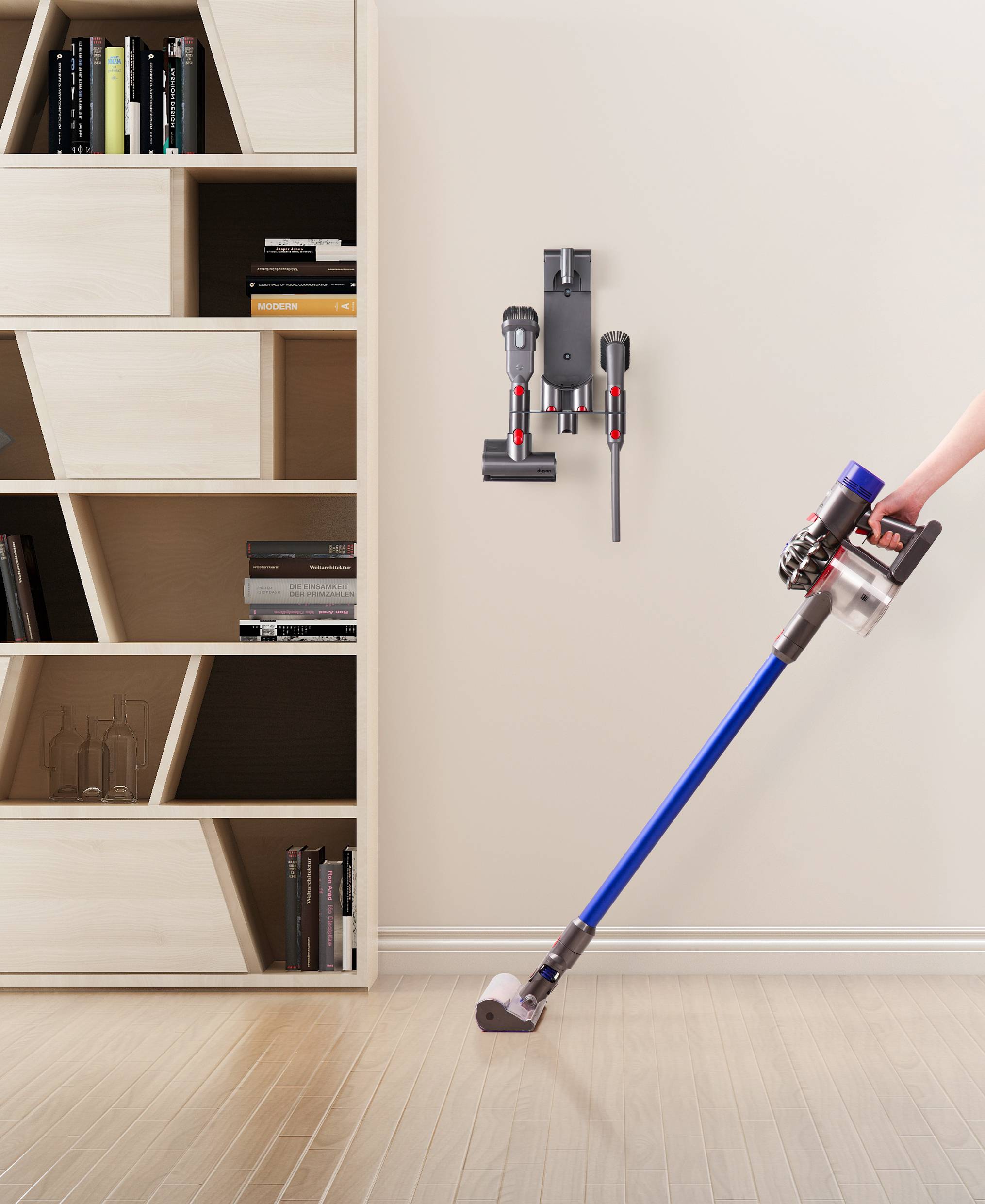 Ein Handstaubsauger wird von einer Person auf einem Holzboden benutzt. An der Wand hängt eine Ladestation mit Zubehör neben einem Regal.