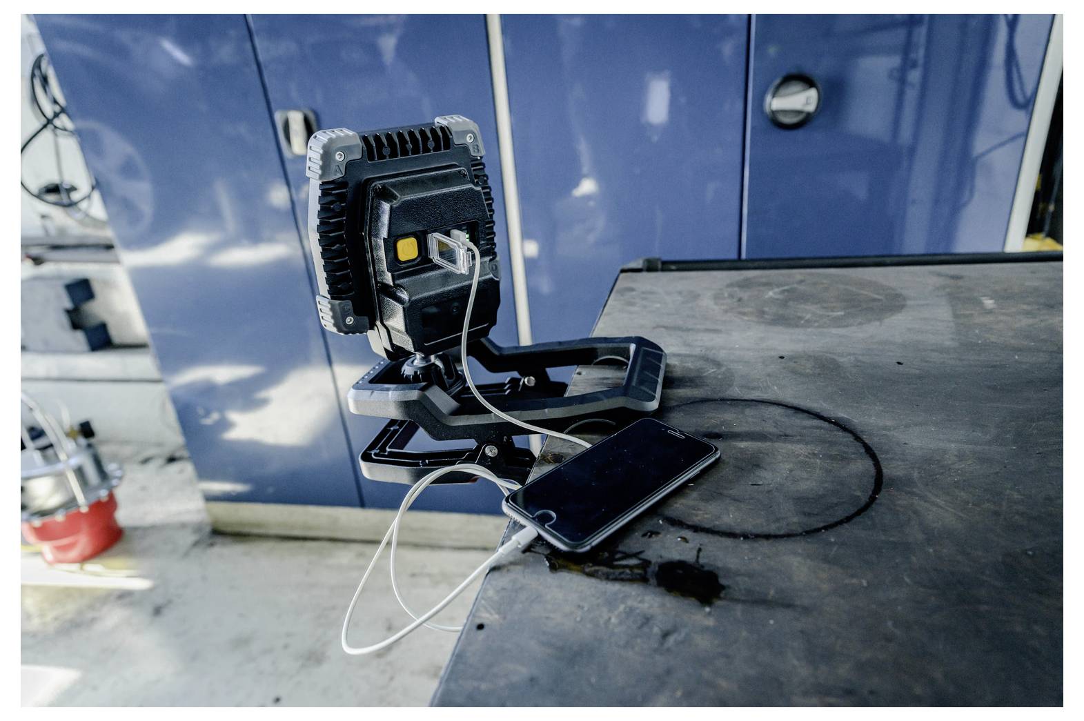 Tragbare Arbeitsleuchte auf einem Tisch mit einem per Kabel verbundenen Smartphone in einer Industriewerkstatt.