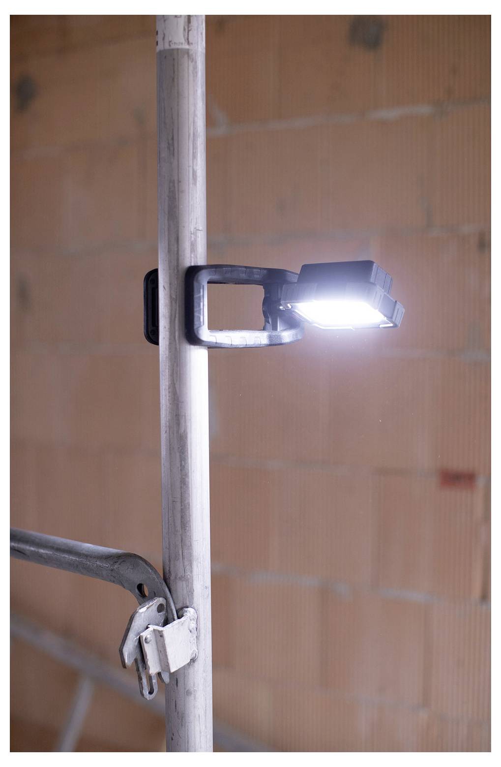 LED-Arbeitsleuchte mit Klemmvorrichtung an einem vertikalen Metallpfosten in einem unfertigen Backsteininterior, mit einer zweiten sichtbaren Klemmvorrichtung darunter.