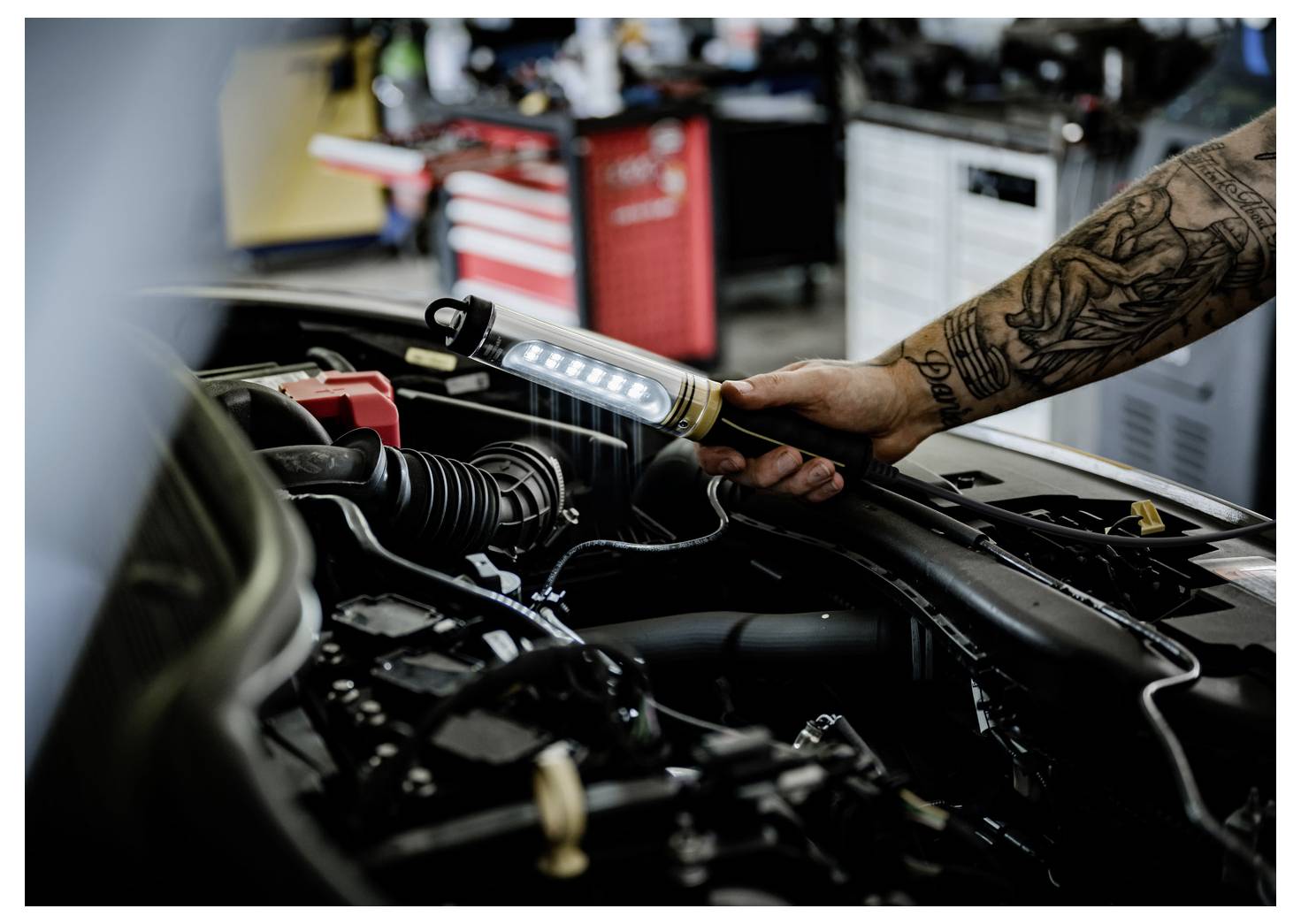 Tätowierter Mechaniker nutzt eine tragbare Arbeitsleuchte, um einen offenen Automotor in einer Werkstatt zu inspizieren.