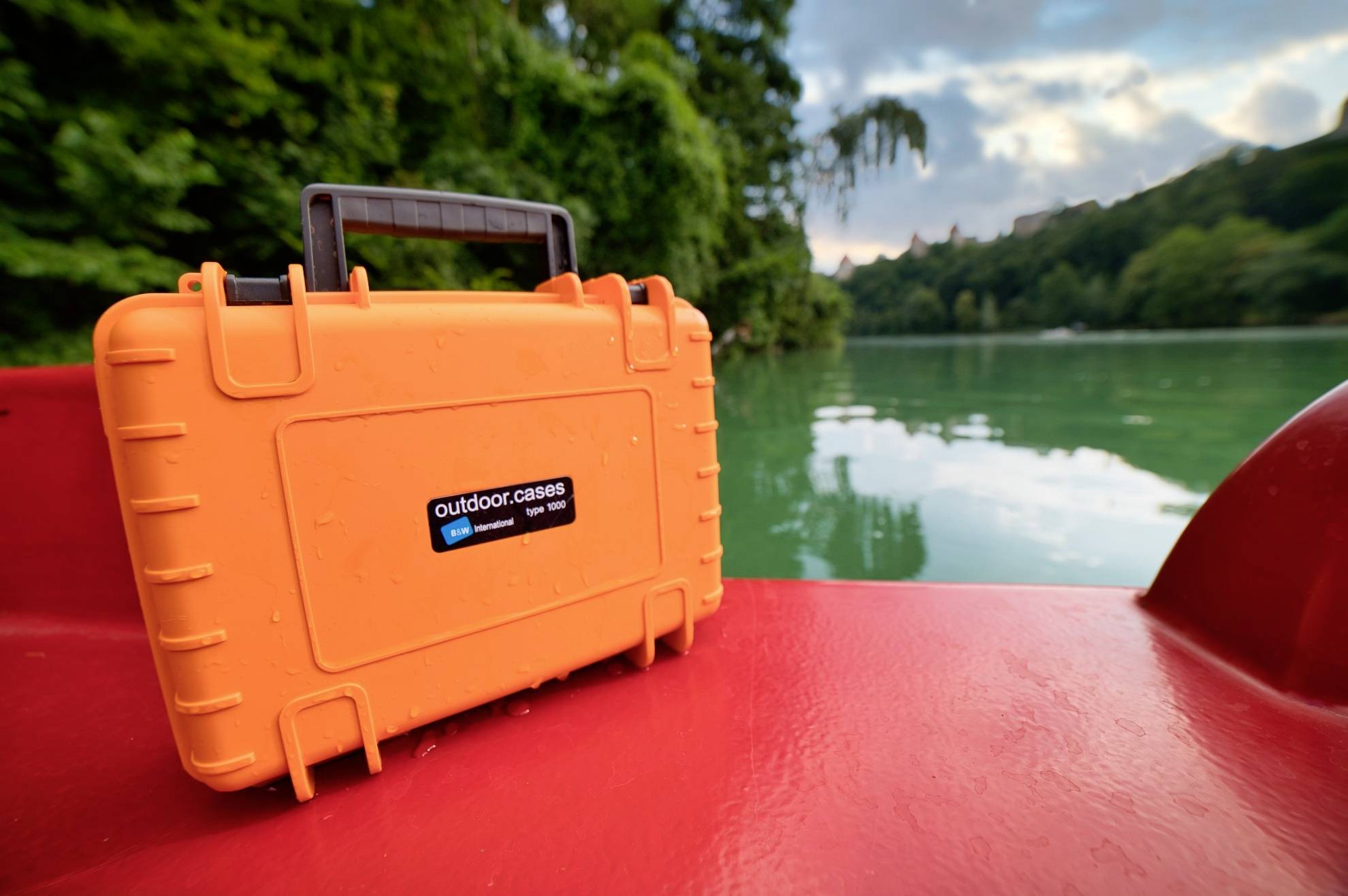 Wasserdichter orangefarbener Koffer mit schwarzem Griff liegt auf rotem Boot in malerischer Flusslandschaft.