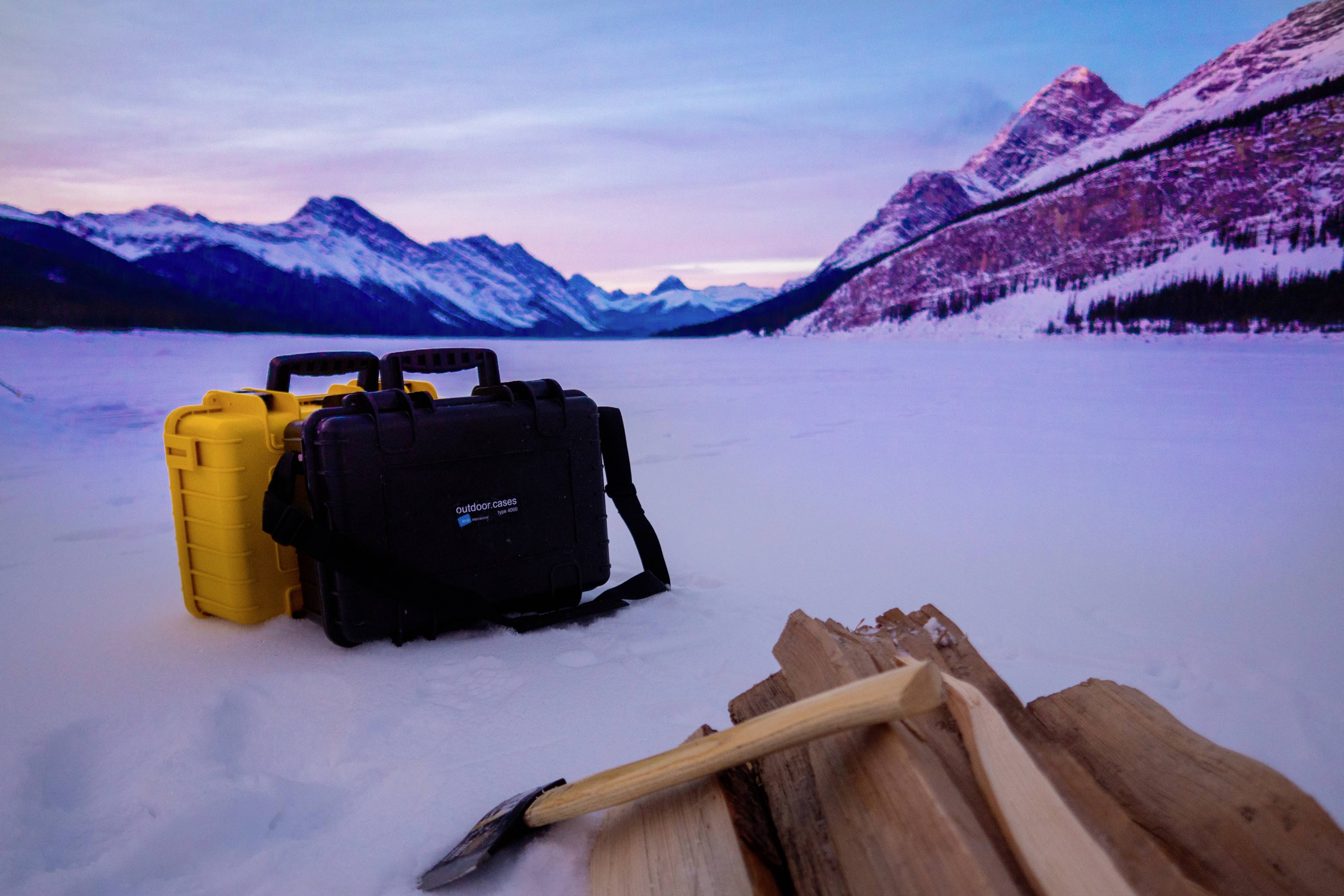 Blick über eine verschneite Gebirgslandschaft bei Dämmerung. Im Vordergrund liegen ein Beil und Holzscheite, daneben zwei Koffer.