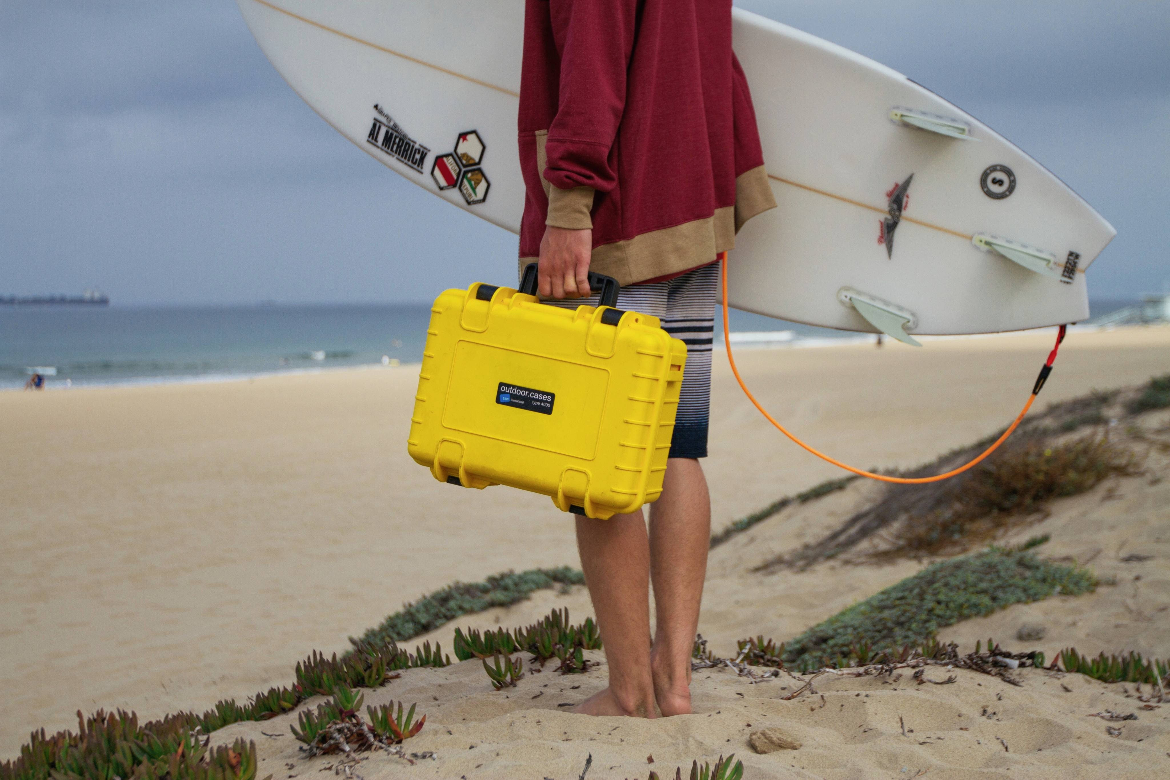 Eine Person steht am Strand mit einem Surfbrett und einem gelben Koffer in der Hand, bereit zum Surfen.