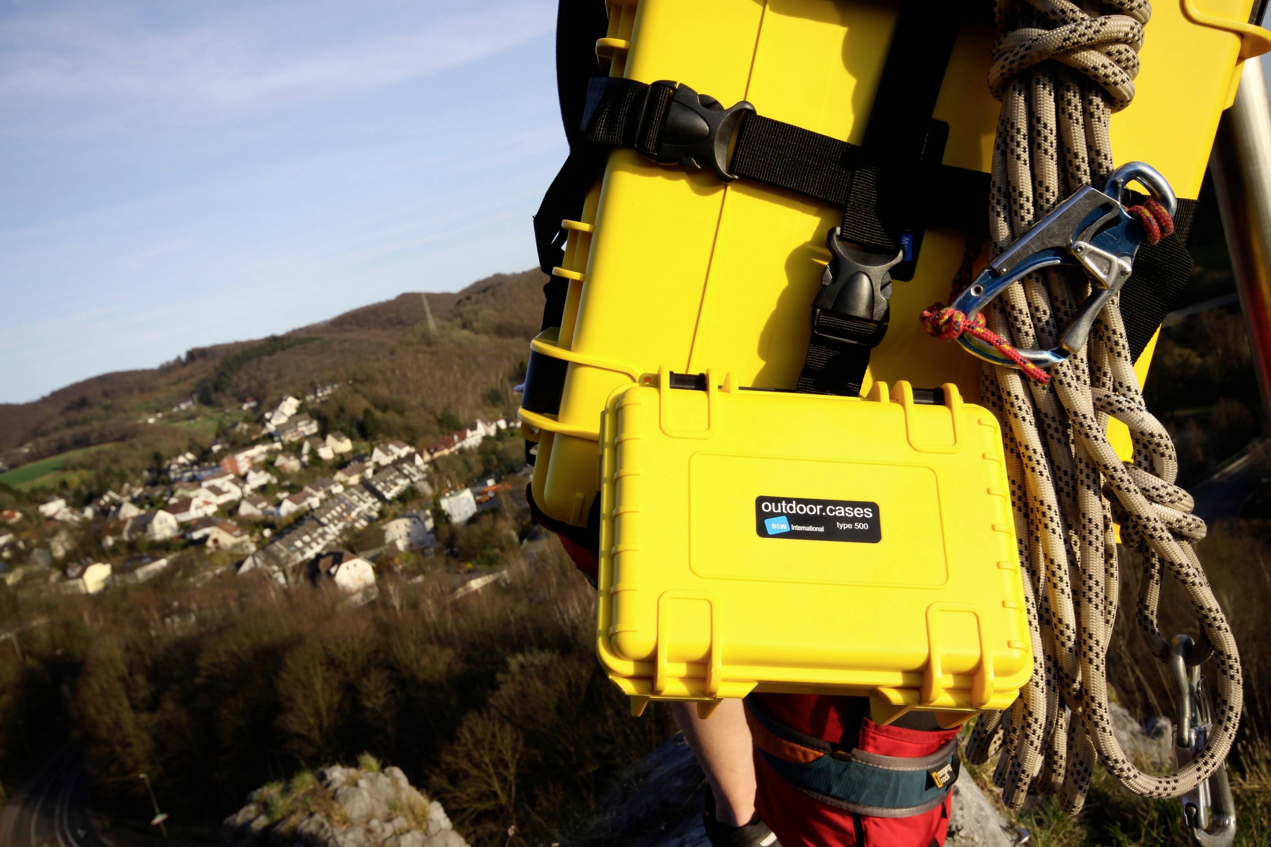 Eine Person trägt zwei gelbe Hartschalenkoffer und ein Seil auf dem Rücken beim Klettern. Im Hintergrund ist eine Landschaft mit Häusern sichtbar.