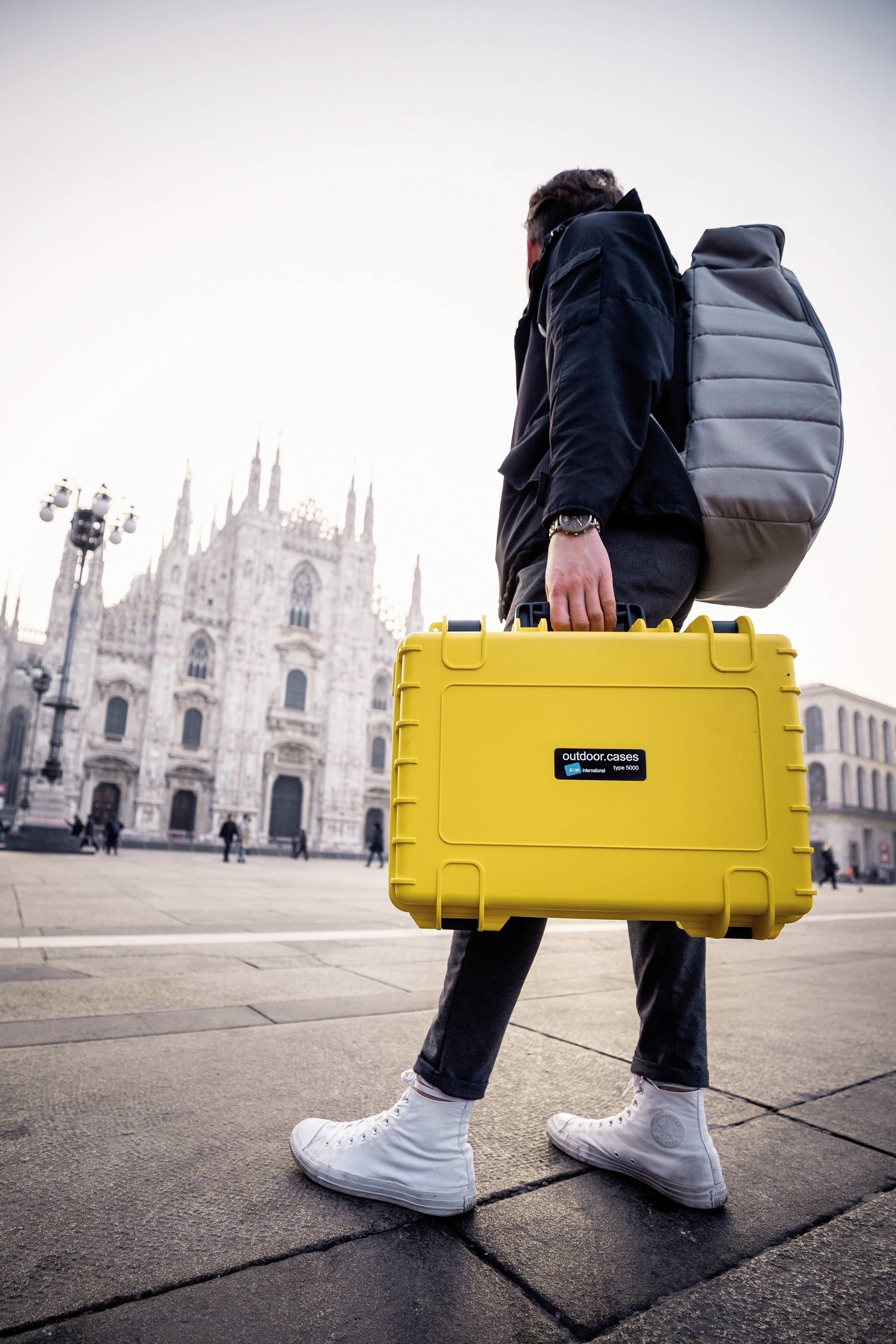 Eine Person in Freizeitkleidung trägt einen gelben Koffer und einen grauen Rucksack vor einer historischen Kathedrale auf einem Platz.