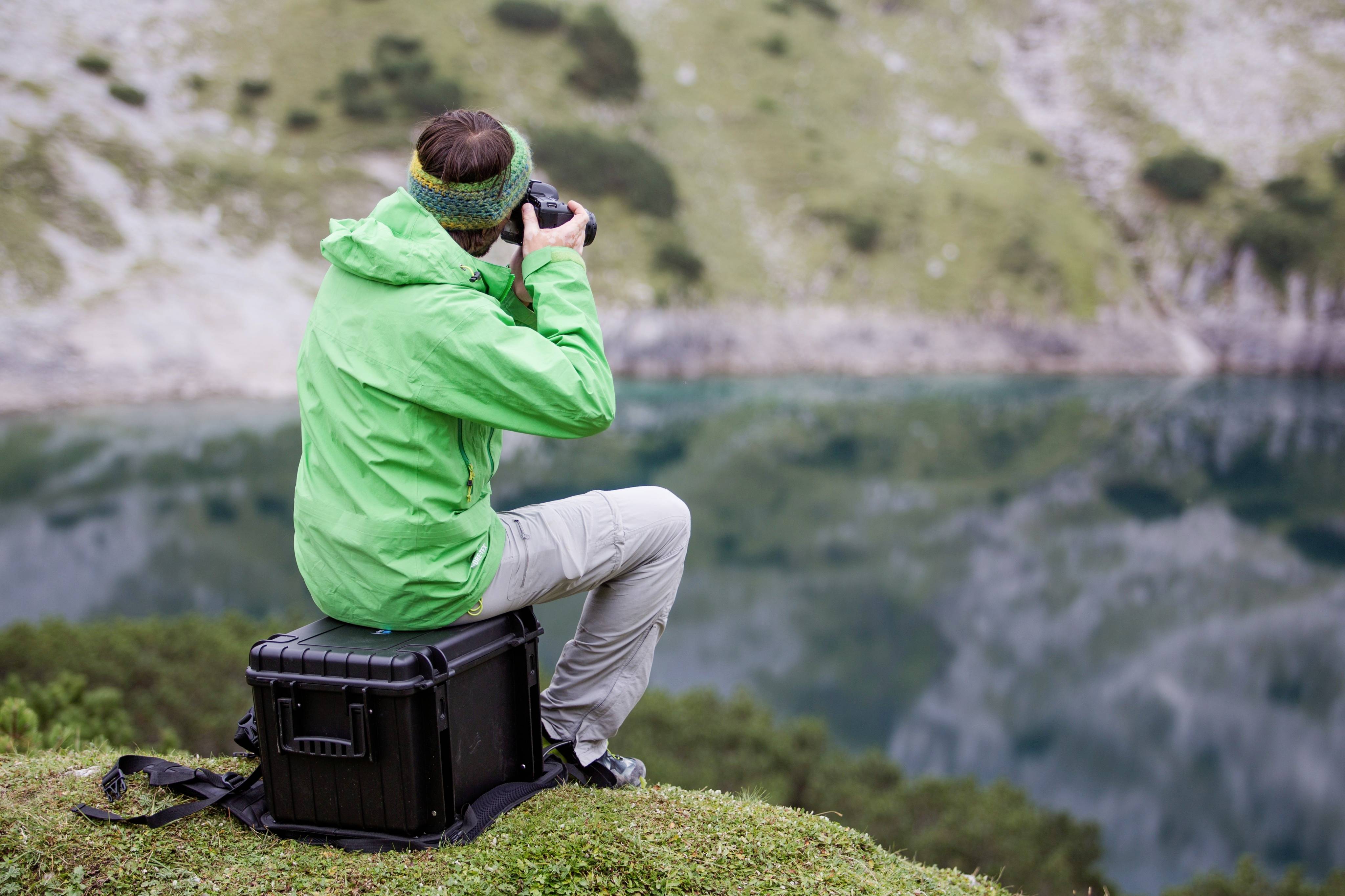 Eine Person in grüner Jacke fotografiert mit einer Kamera auf einem Koffer sitzend, vor einem See in einer bergigen Landschaft.