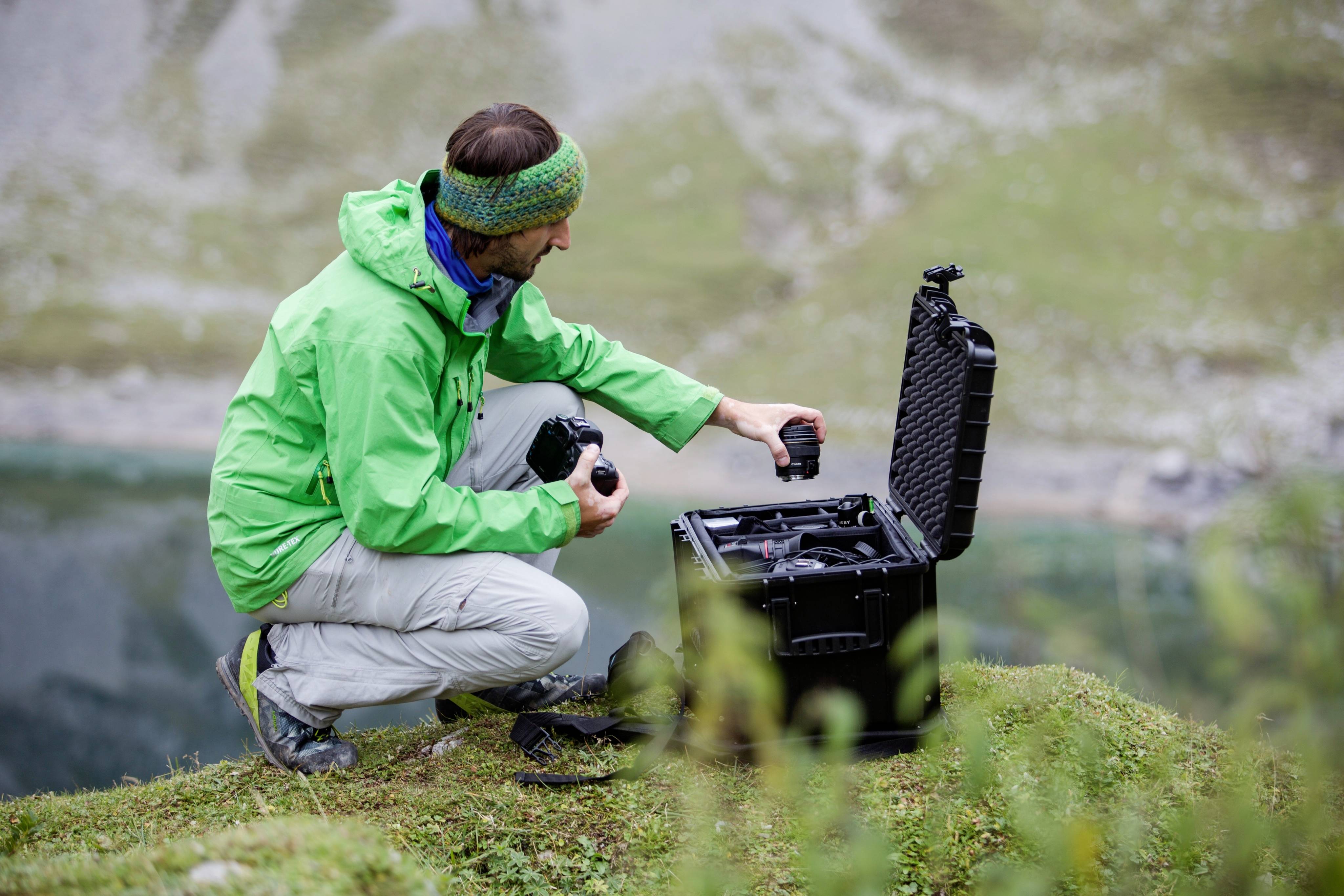 Eine Person in grüner Jacke kniet auf einer Wiese vor einer offenen schwarzen Tasche mit Drohnenausrüstung, umgeben von natürlicher Landschaft.