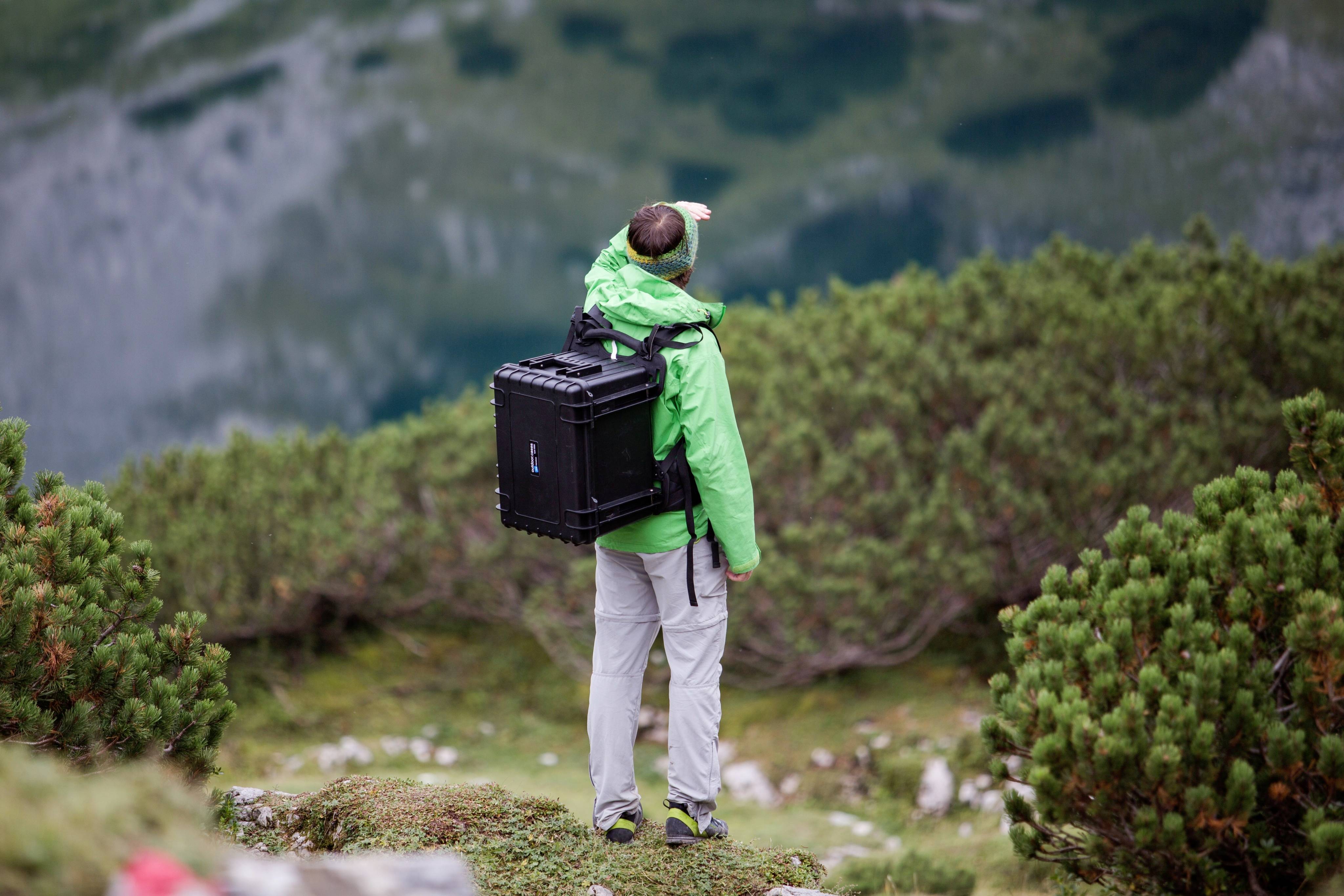 Ein Mann in grüner Jacke steht in alpiner Landschaft mit Rucksack und zeigt auf einen Punkt in der Ferne. Dichte Vegetation umgibt ihn.
